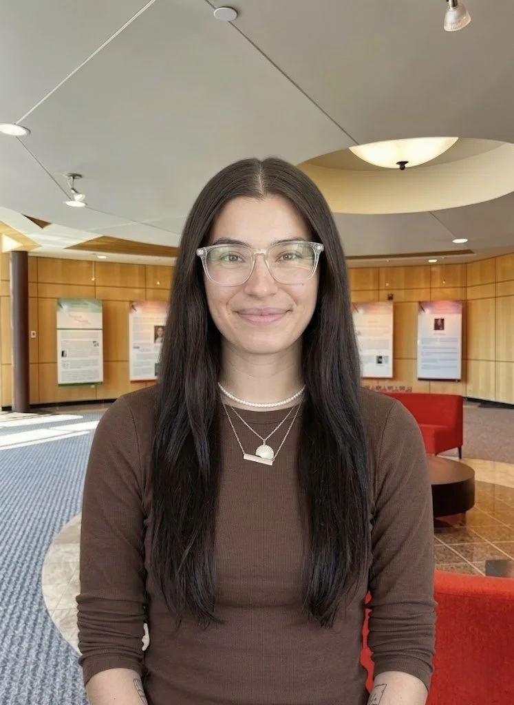 A young woman with curly hair and blue eyes smiling at the camera, wearing a black top with gold buttons on the shoulder, indoors in a warmly lit space.