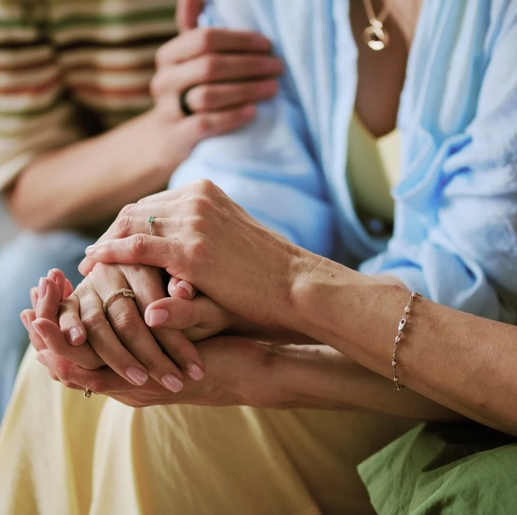 Close-up of two women holding hands, showing wedding rings and jewelry, with one woman clad in a light blue shirt and the other in a striped top.