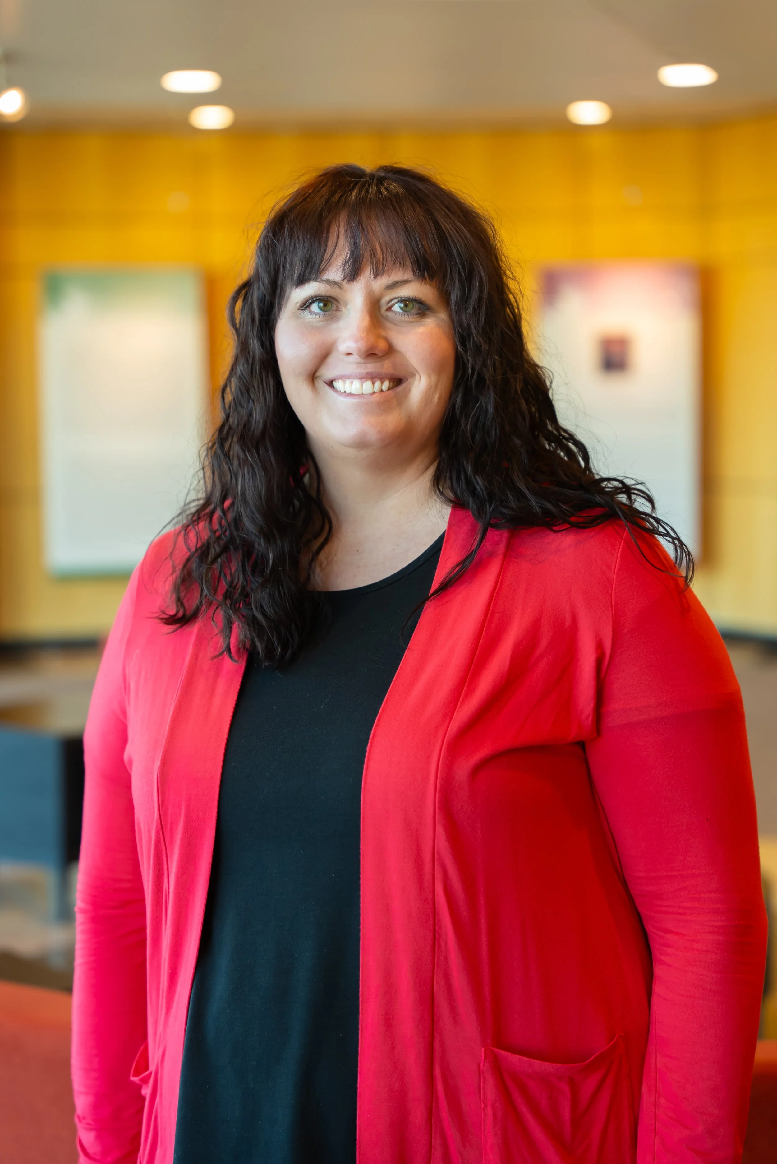 A woman with dark brown, curly hair wearing a red blazer over a black shirt, smiling indoors with a yellow wall and blurred framed pictures in the background.