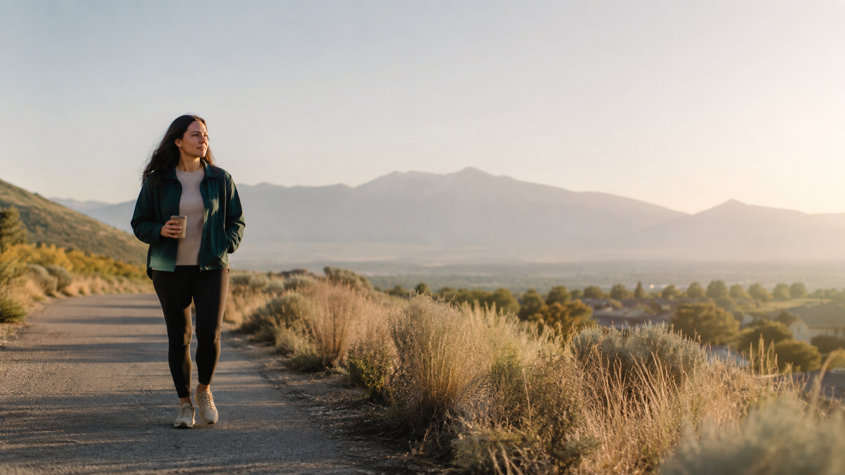 A woman walking on a dirt path holding a coffee cup, with mountains and a valley in the background during sunset.