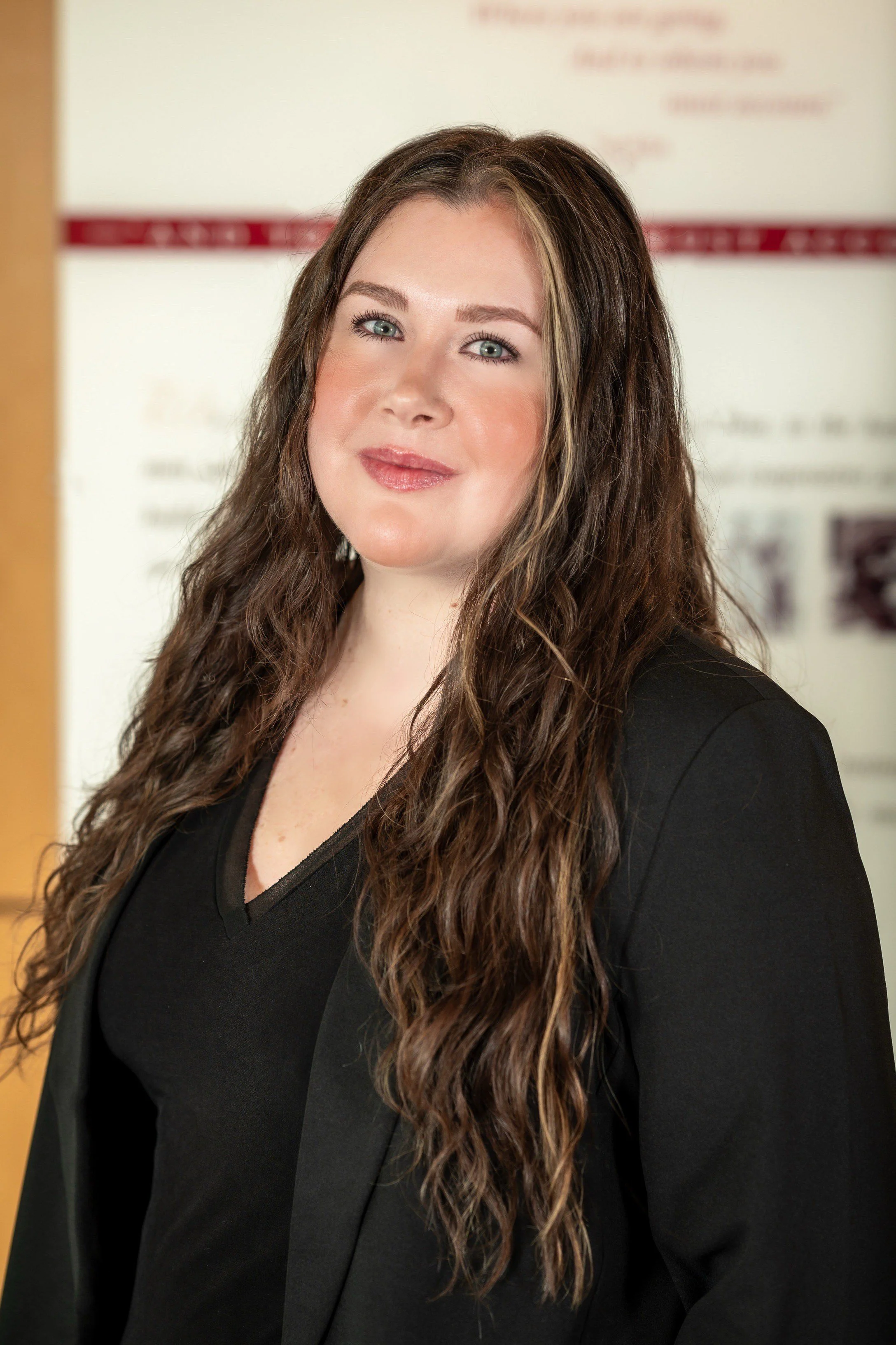 A woman with long, curly brown hair and blue eyes, wearing a black blazer over a black top, standing indoors near a bulletin board or wall with posters or papers.