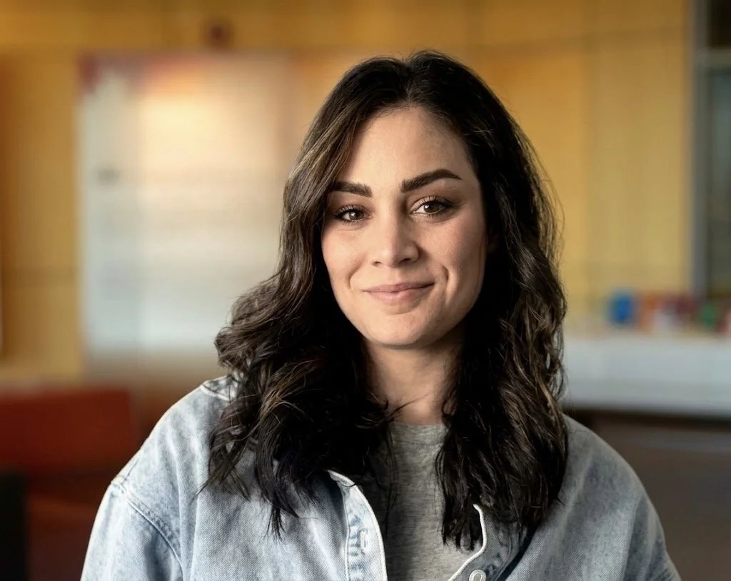 A young woman with dark wavy hair smiling in an indoor setting with warm lighting.
