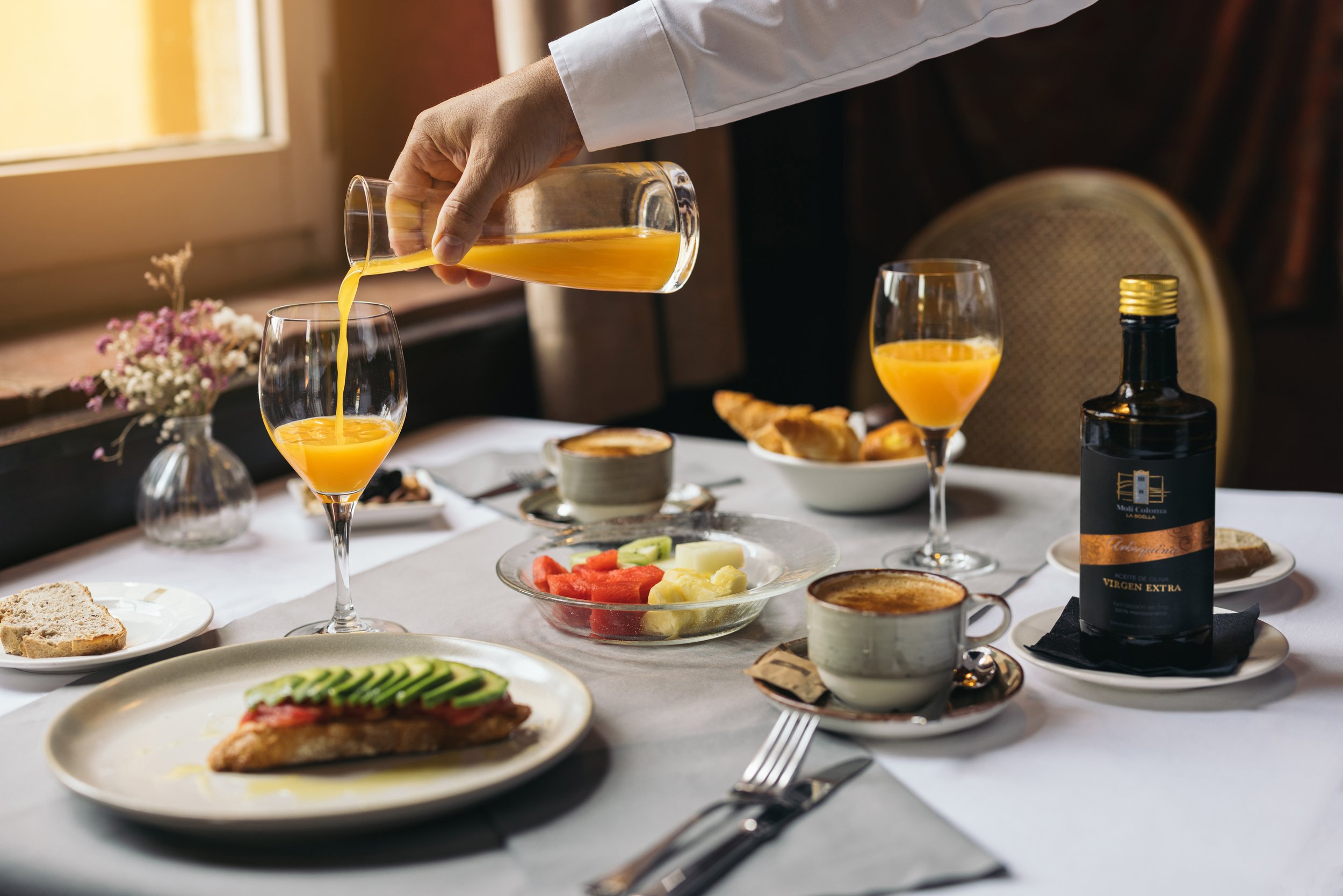 Mesa de desayuno con jugo de naranja, fruta, café y pan, en un ambiente acogedor