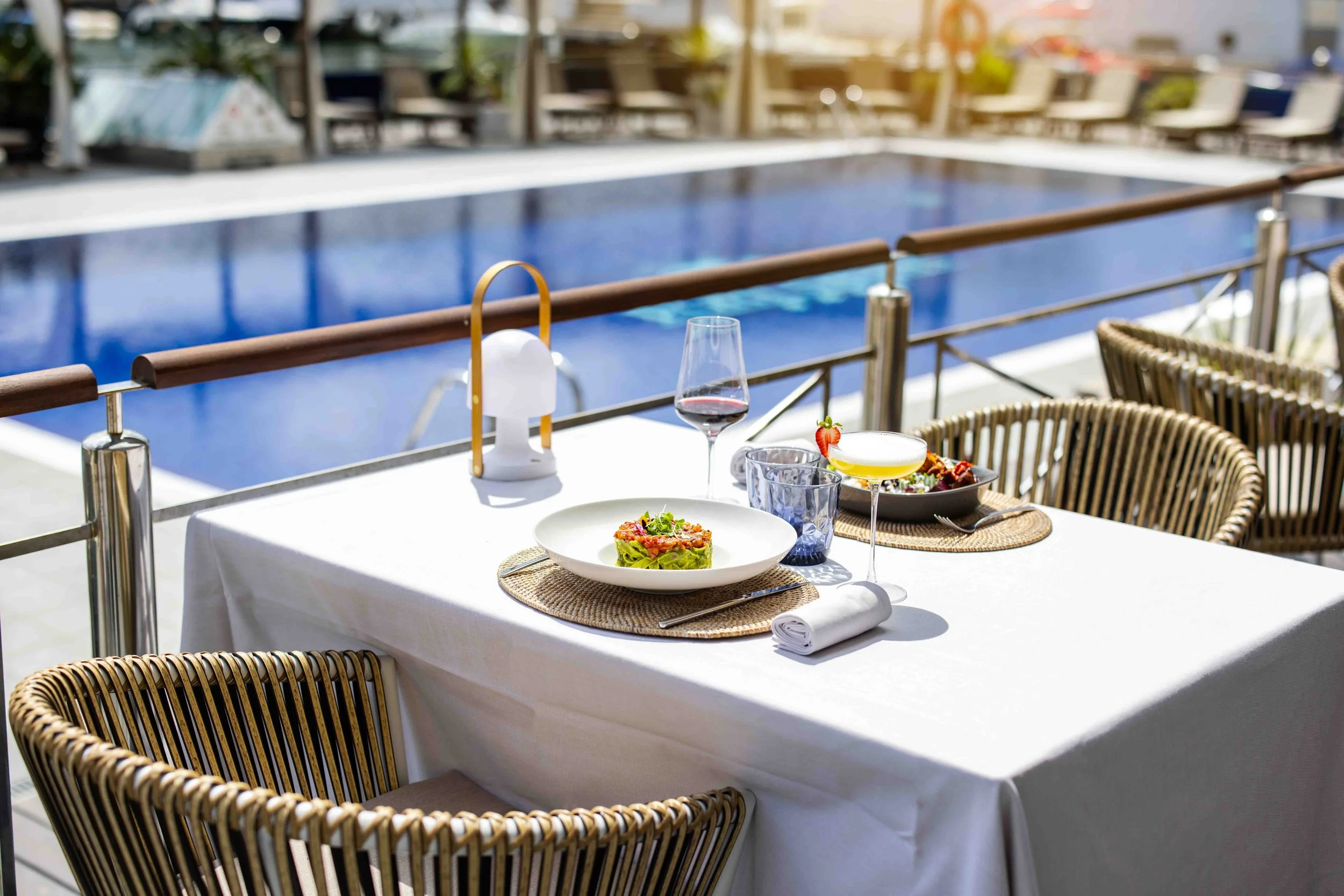 Mesa puesta con platos de comida, copas de vino y agua, en un área junto a una piscina.