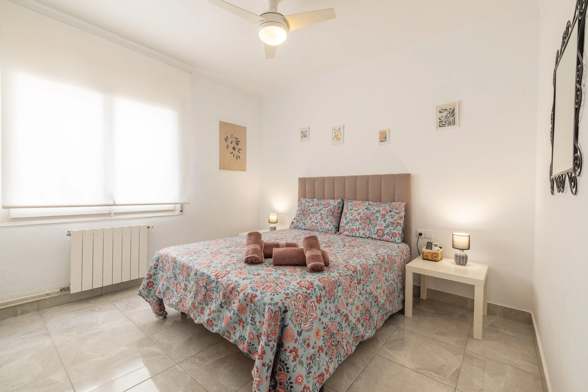 Habitación con cama de matrimonio, sábanas con diseño floral, acolchado beige, paredes blancas, ventana con persiana, lámparas de mesa, cuadros decorativos, espejo y aire acondicionado