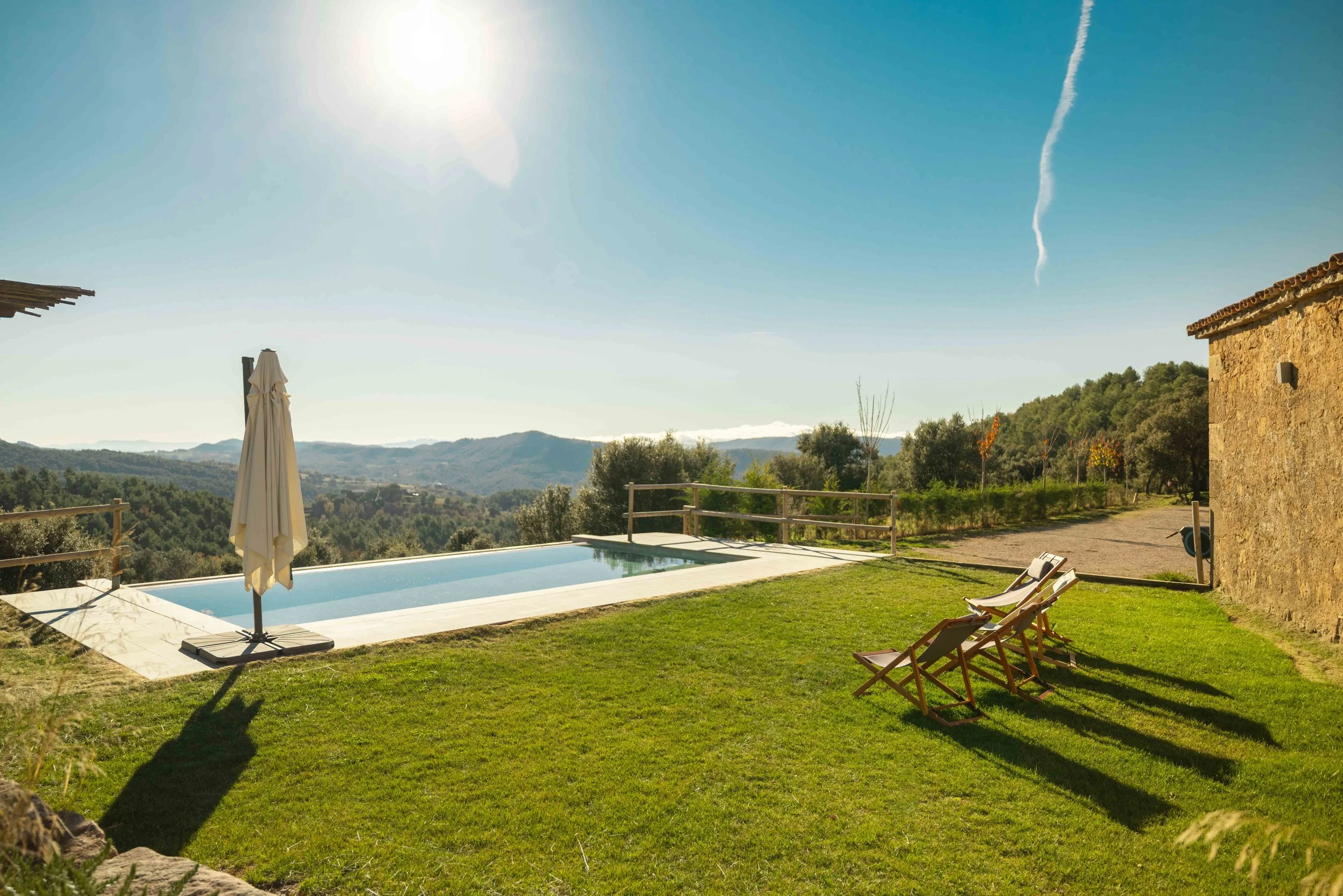 Piscina rectangular con vista a las colinas, sombrilla cerrada y dos sillas de lona en césped, cerca de una casa de piedra, en un día soleado con cielo despejado.