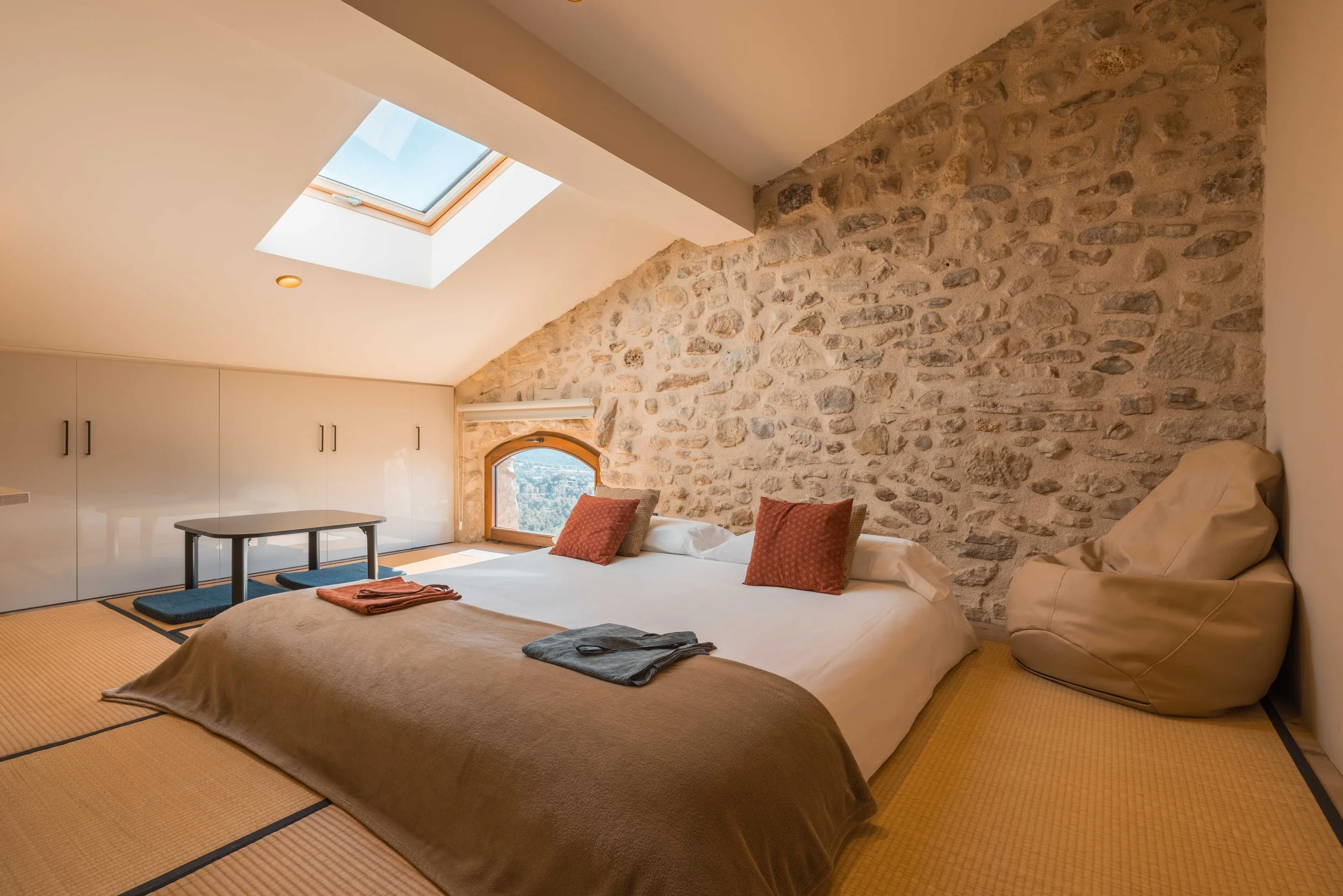 Habitación con techo inclinado, pared de piedra, cama grande con almohadas y toallas, ventana pequeña con vista a la naturaleza, luz natural y decoración cálida.
