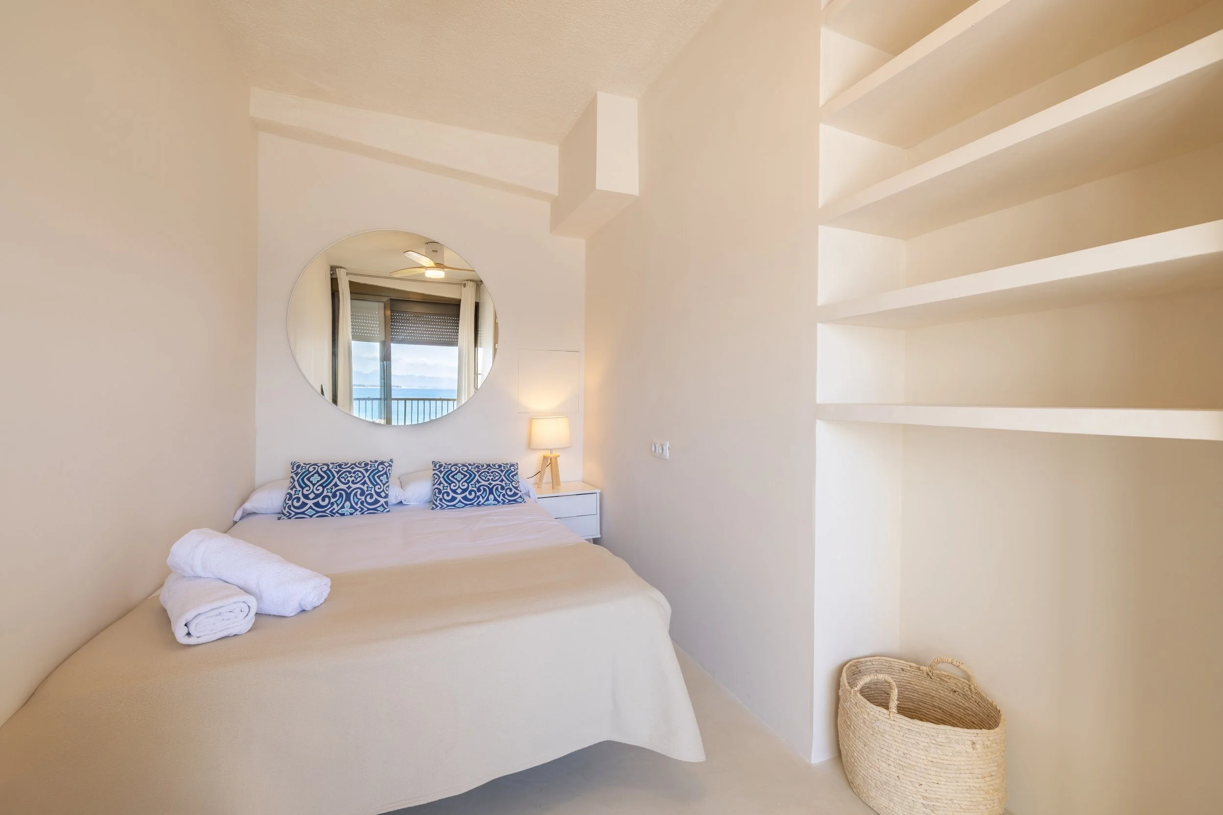 Habitación sencilla con cama, almohadas azules, espejo redondo en la pared, lámpara junto a la cama, ventanas con vista al mar.