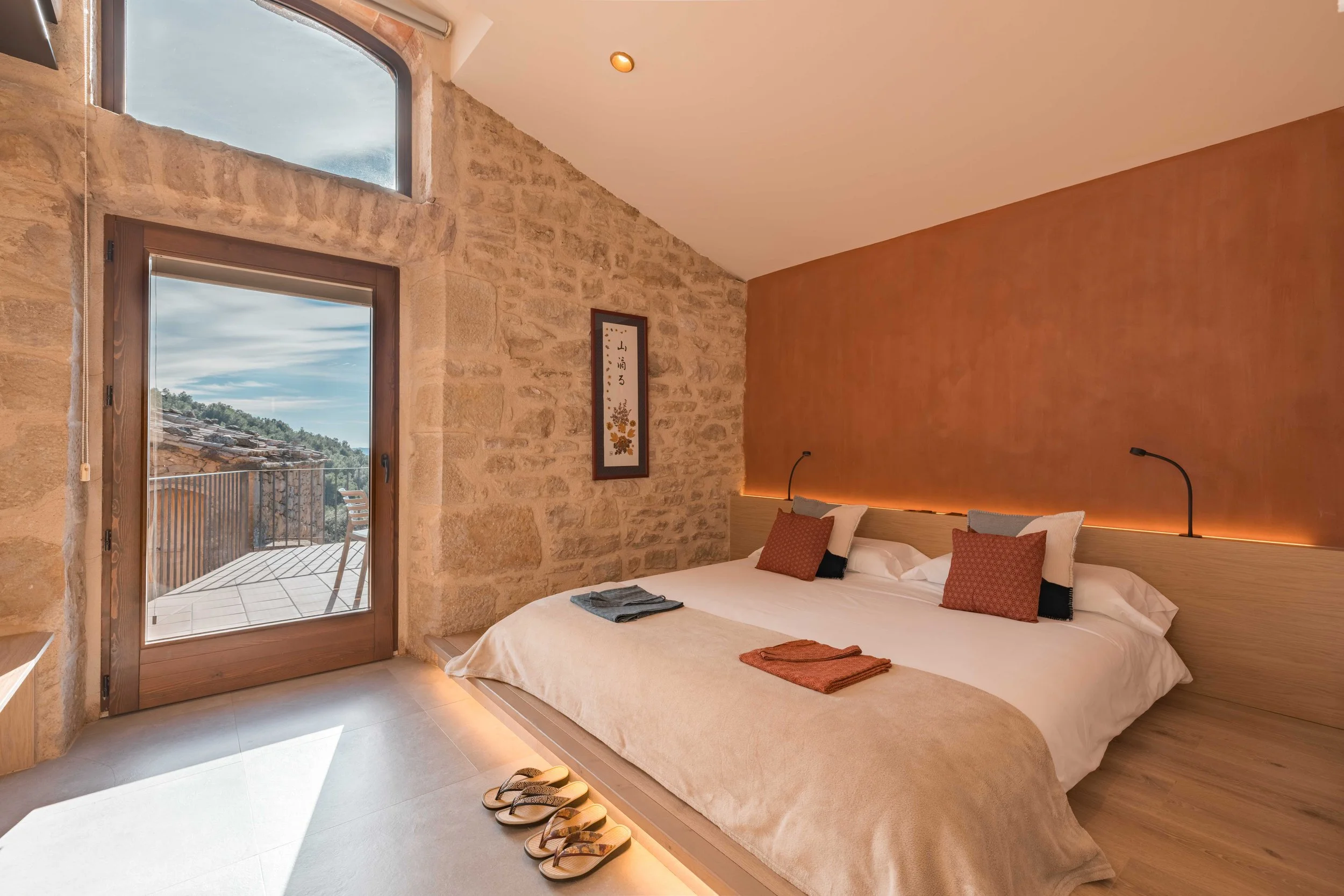 Habitación moderna con paredes de piedra, cama grande con almohadas blancas y naranjas, vista a un balcón con paisaje montañoso, ilustración artística en la pared.