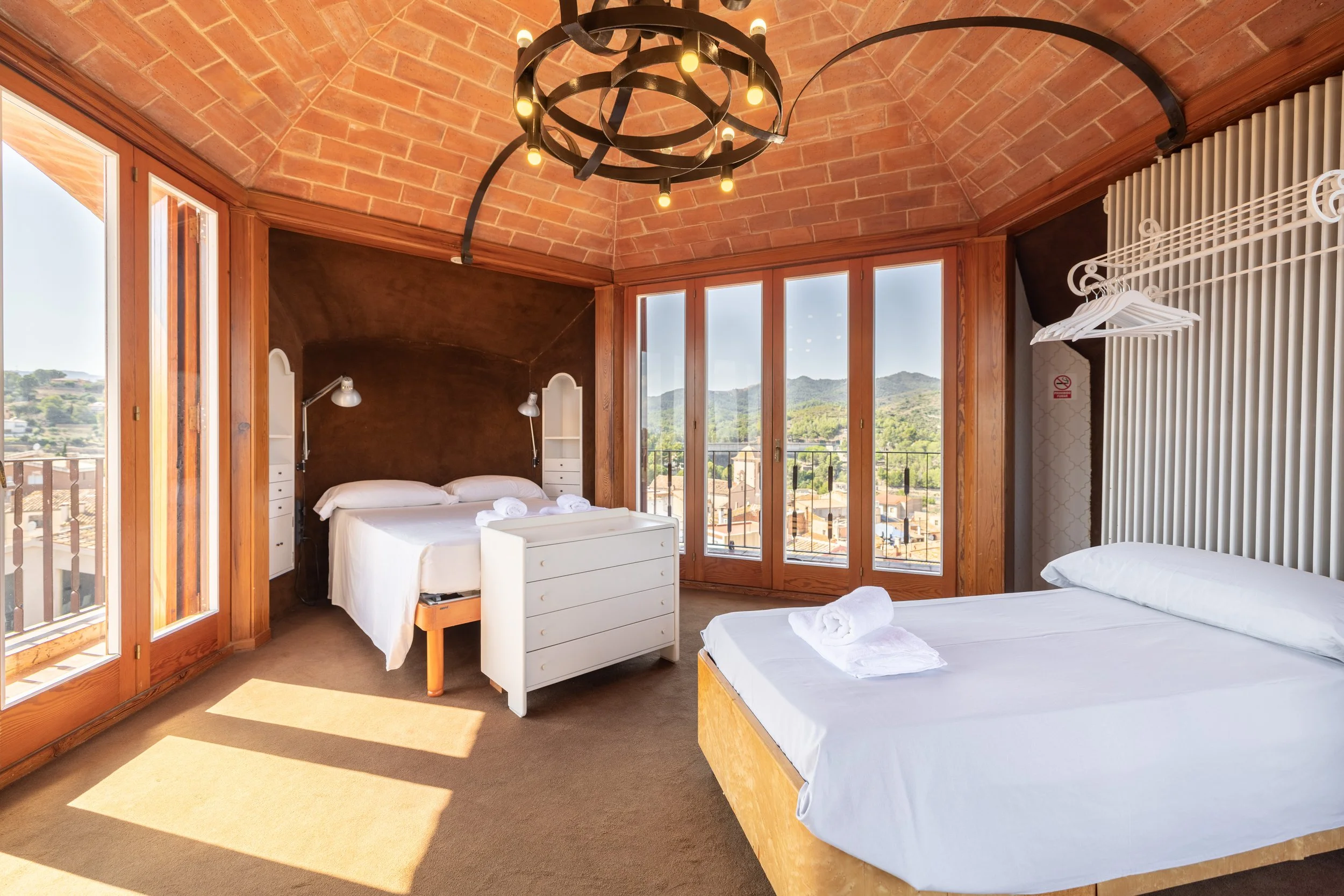 Habitación con dos camas de matrimonio, paredes de ladrillo, ventanas grandes con vista a las montañas, lámpara de techo y mobiliario de madera.