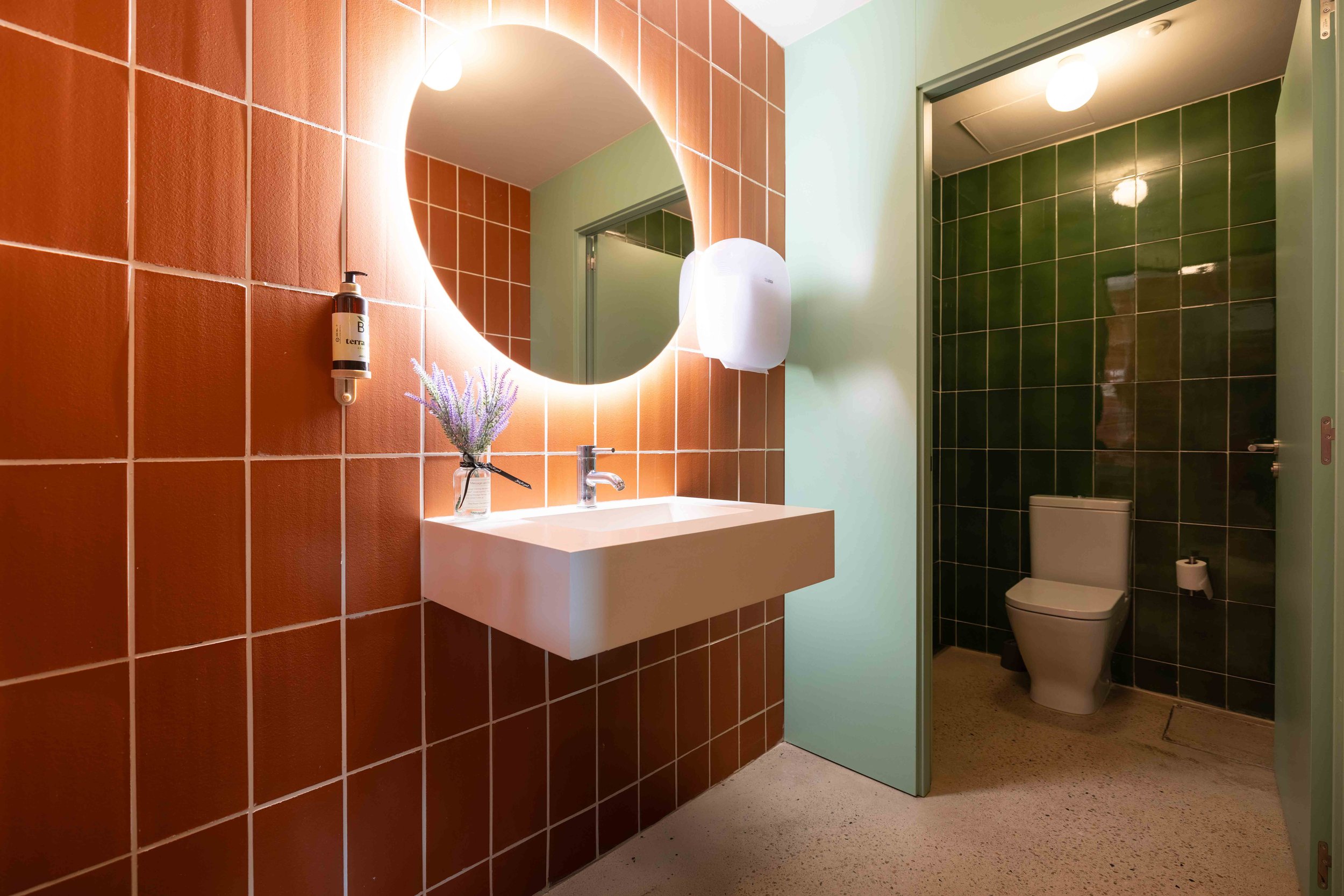 Baño con pared de azulejos rojos, lavabo blanco con espejo redondo iluminado, planta decorativa, dispensador de jabón, y váter en un espacio separado con paredes de azulejos verdes.