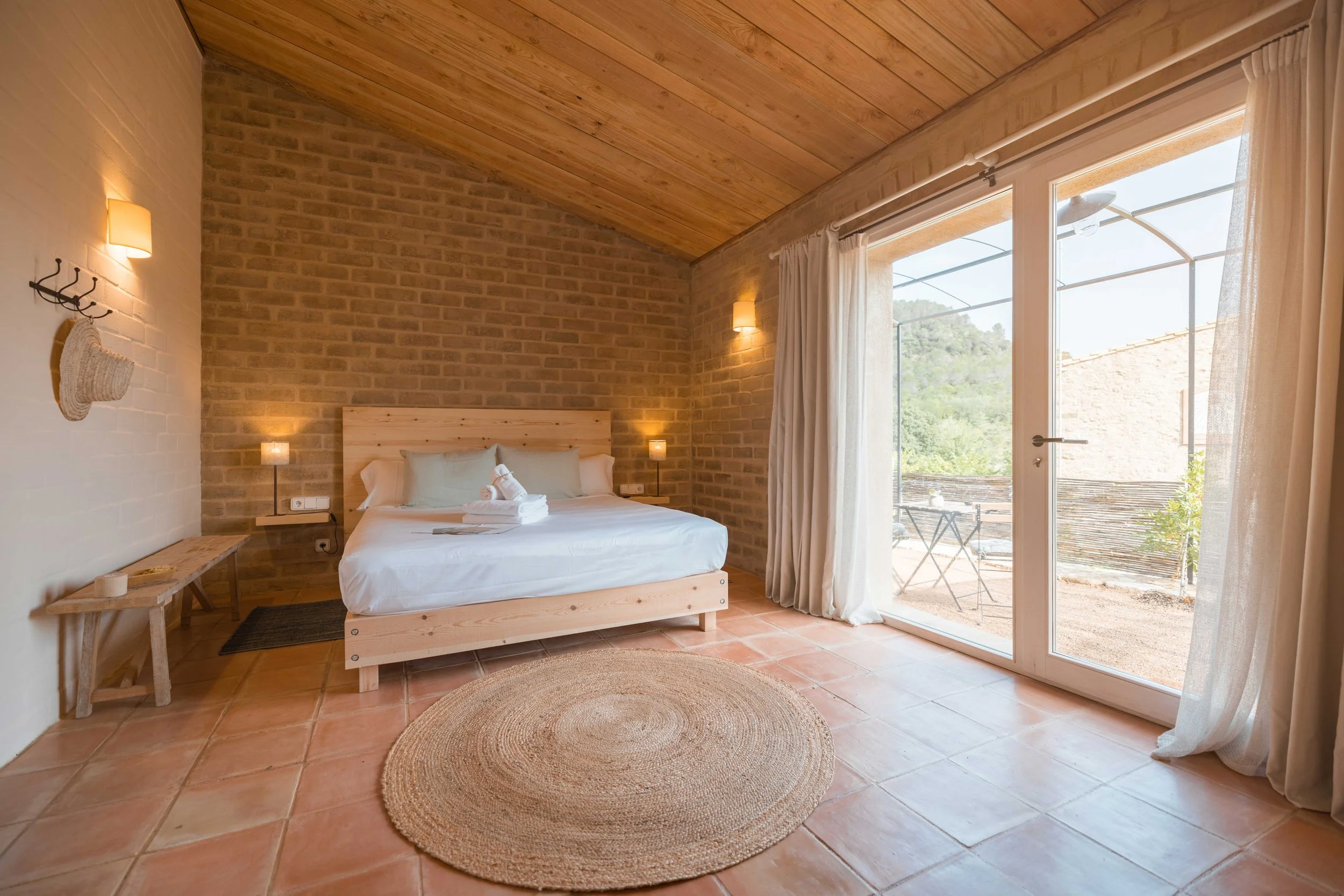 Habitación con cama doble, paredes de ladrillo visible, techo de madera, piso de baldosas, cortinas blancas, ventana grande con vista a un patio con sillas y mesa, decoración sencilla con lámparas y una alfombra redonda de fibra natural.