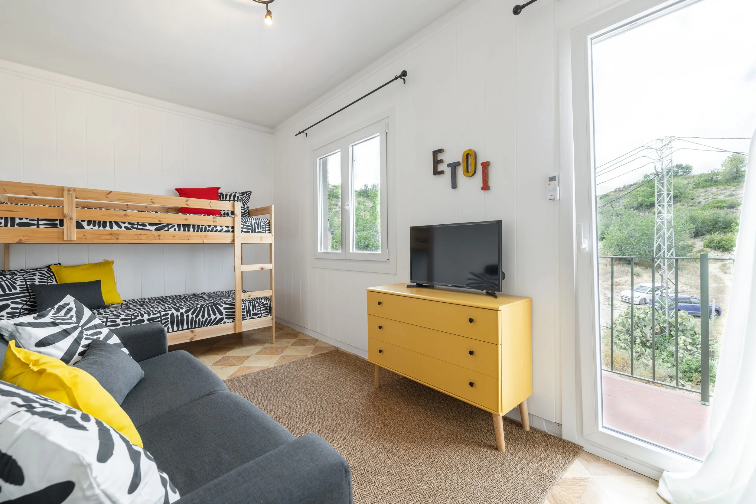 Sala de estar con sofá gris, almohadas amarillas y negras, cama con literas de madera, vista a un balcón con plantas y autos, decoraciones en letras coloridas en la pared.