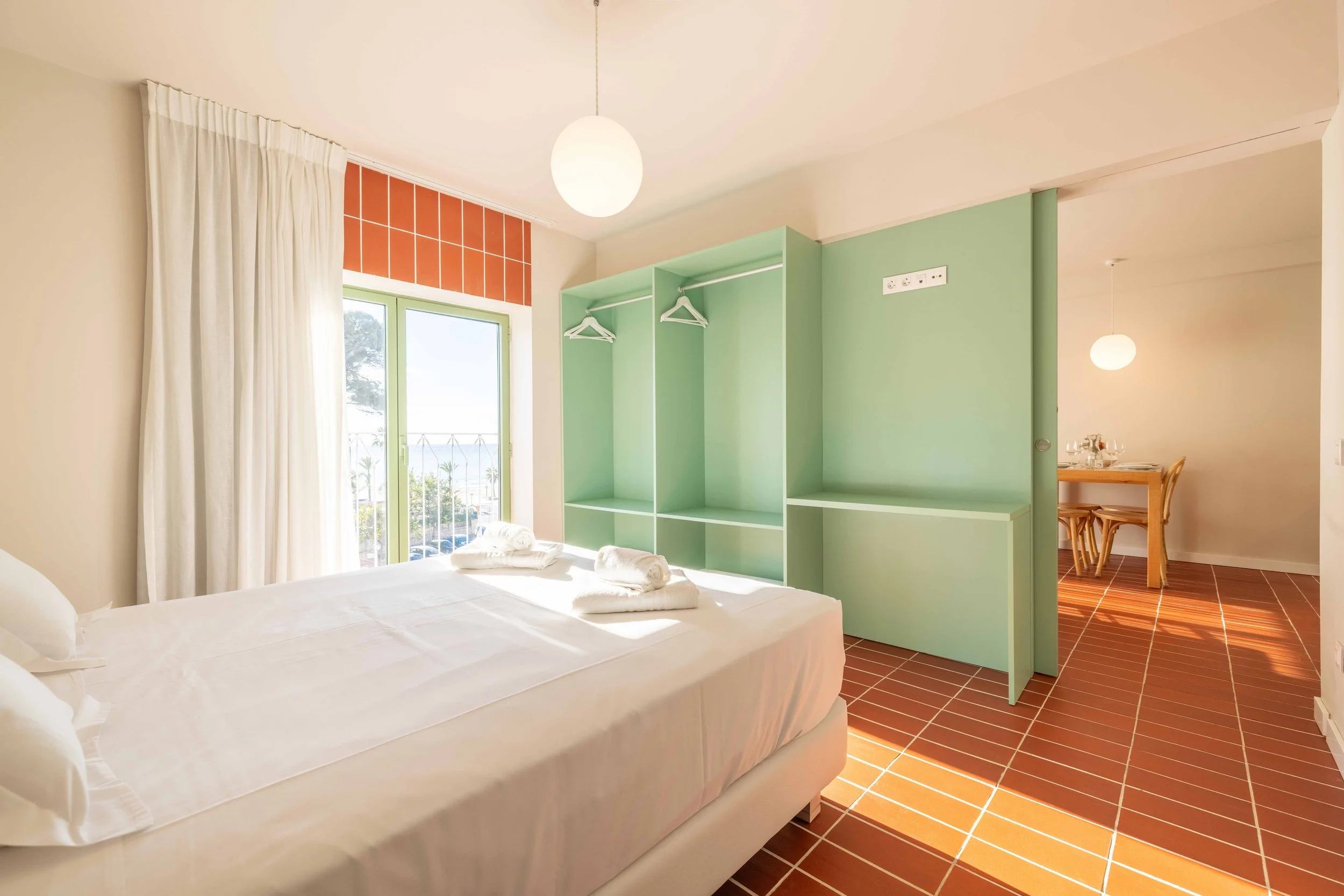 Habitación con cama doble, ventanas con cortinas blancas, muebles verdes, y un comedor en el fondo, con suelo de baldosas terracota.