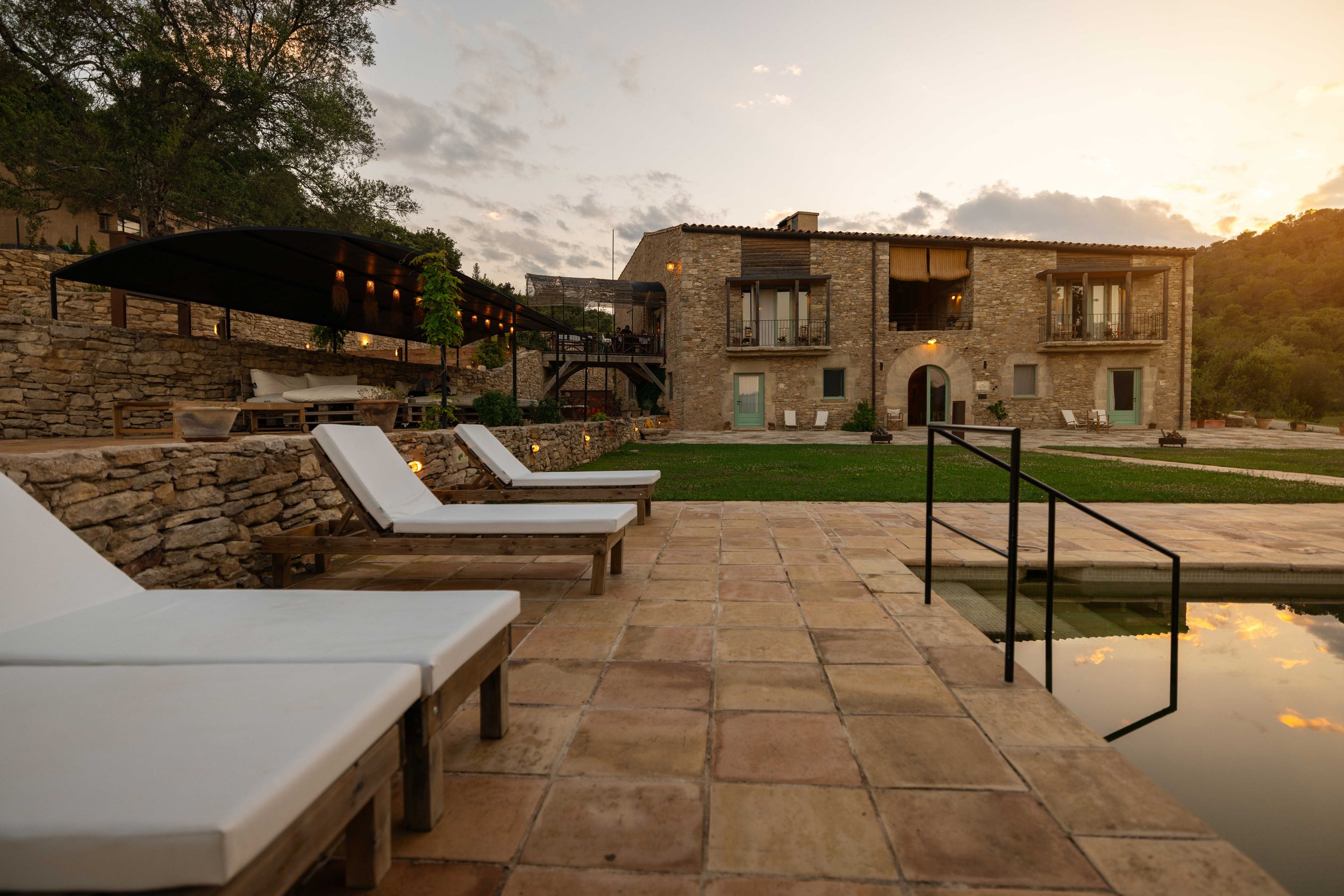 Vista de una casa de piedra al atardecer con tumbonas blancas, piscina y jardín.