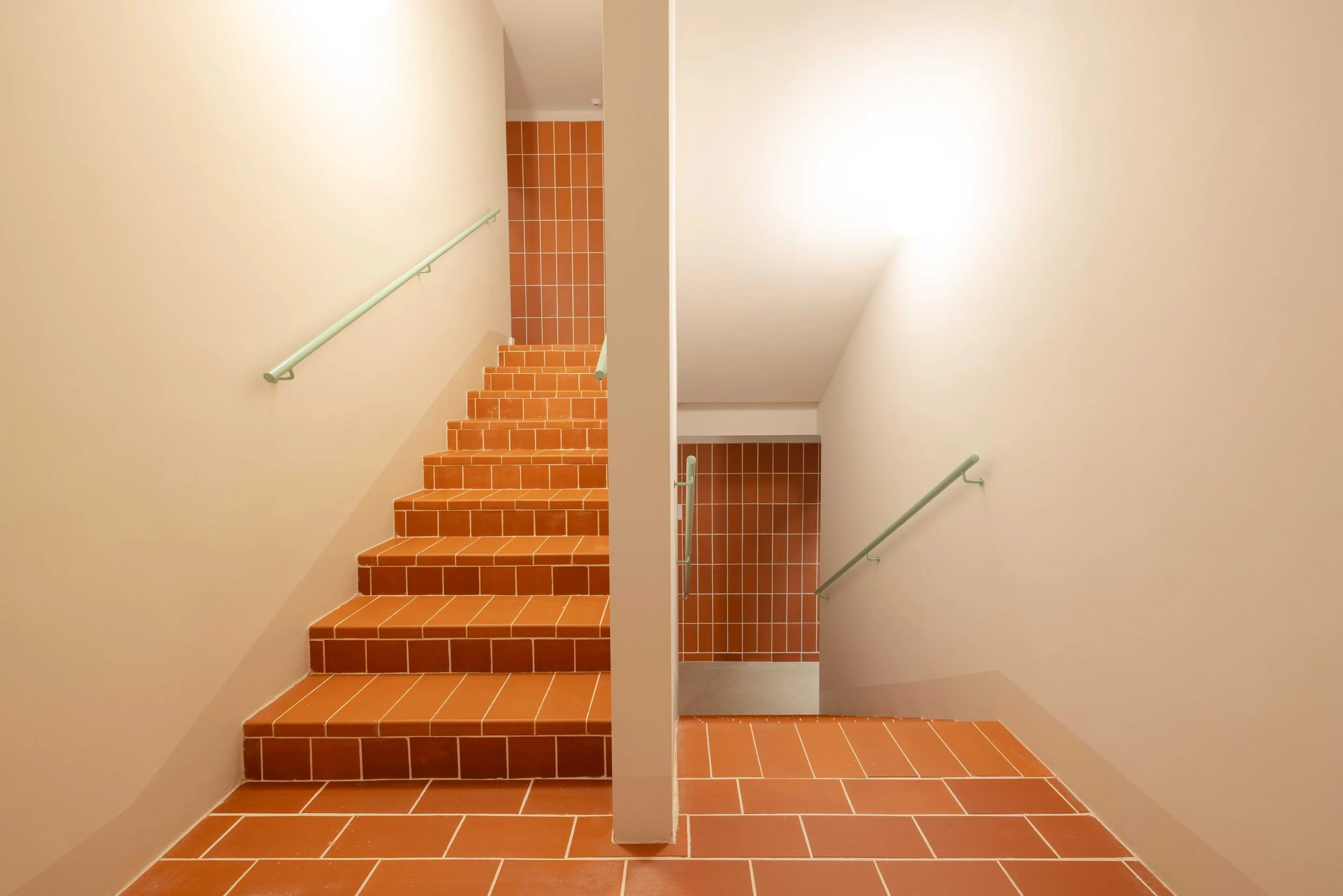 Escaleras interiores con barandillas de color verde y paredes blancas, suelo y parte de la pared de fondo con baldosas de color terracota.
