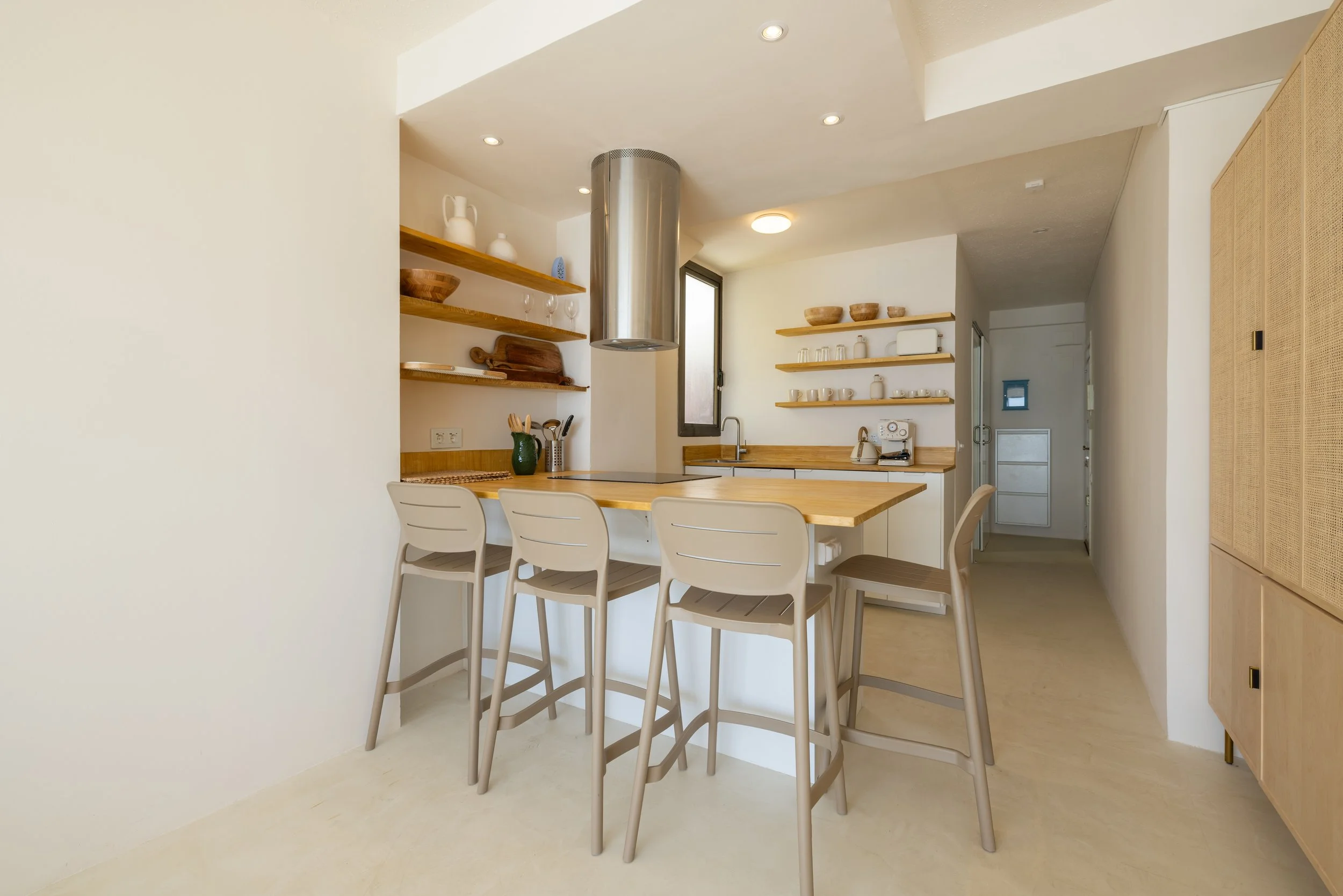 Cocina moderna con barra de madera, cinco sillas, estantes con platos y utensilios, fregadero, ventana, y espacio limpio y luminoso.