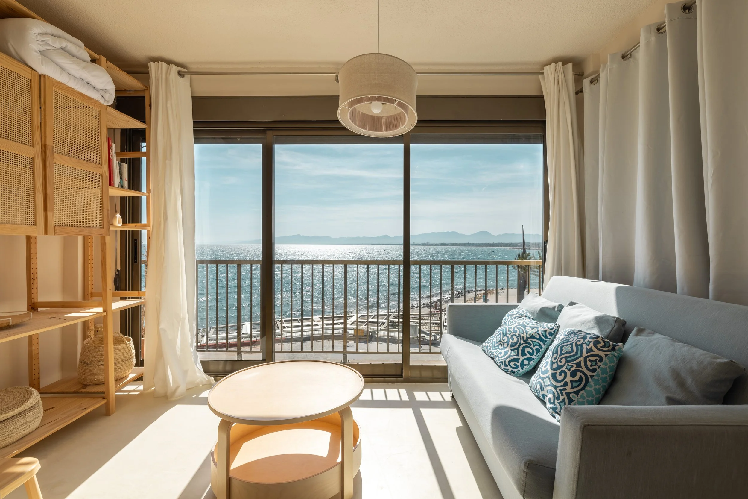 Sala de estar con vista al mar, sofá gris con cojines decorativos, estantería de madera, mesa de centro, cortinas blancas y balcón con barandilla frente al océano