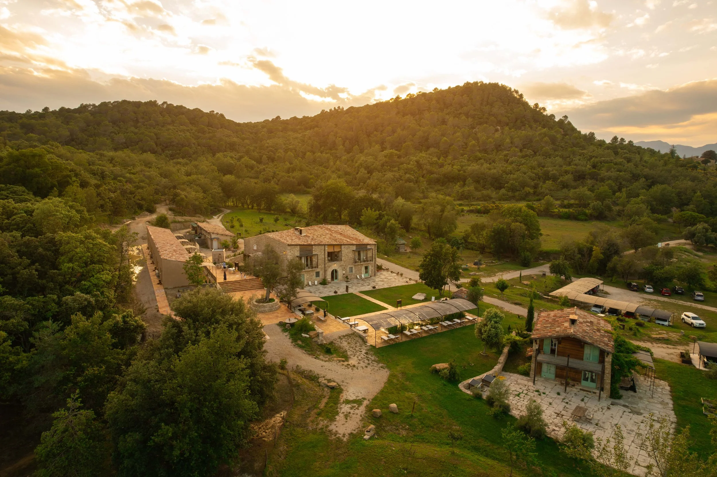 Fotografía de vista panorámica de una finca en una zona montañosa con árboles, edificios rústicos y caminos en un atardecer con cielo nublado.