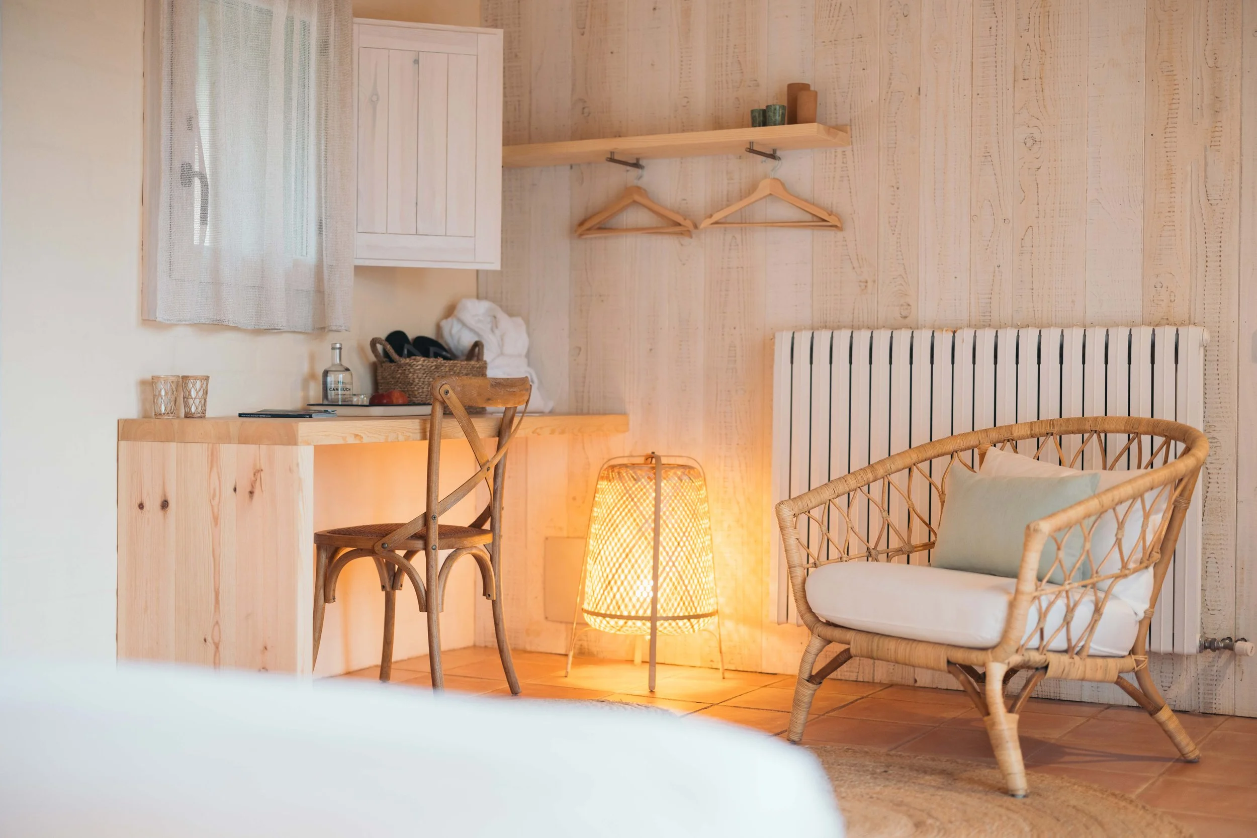 Rincón de habitación con mobiliario de madera clara, sillón de mimbre con cojín, lámpara de pie encendida, escritorio con silla de madera, estanterías con objetos decorativos y paredes de madera clara.