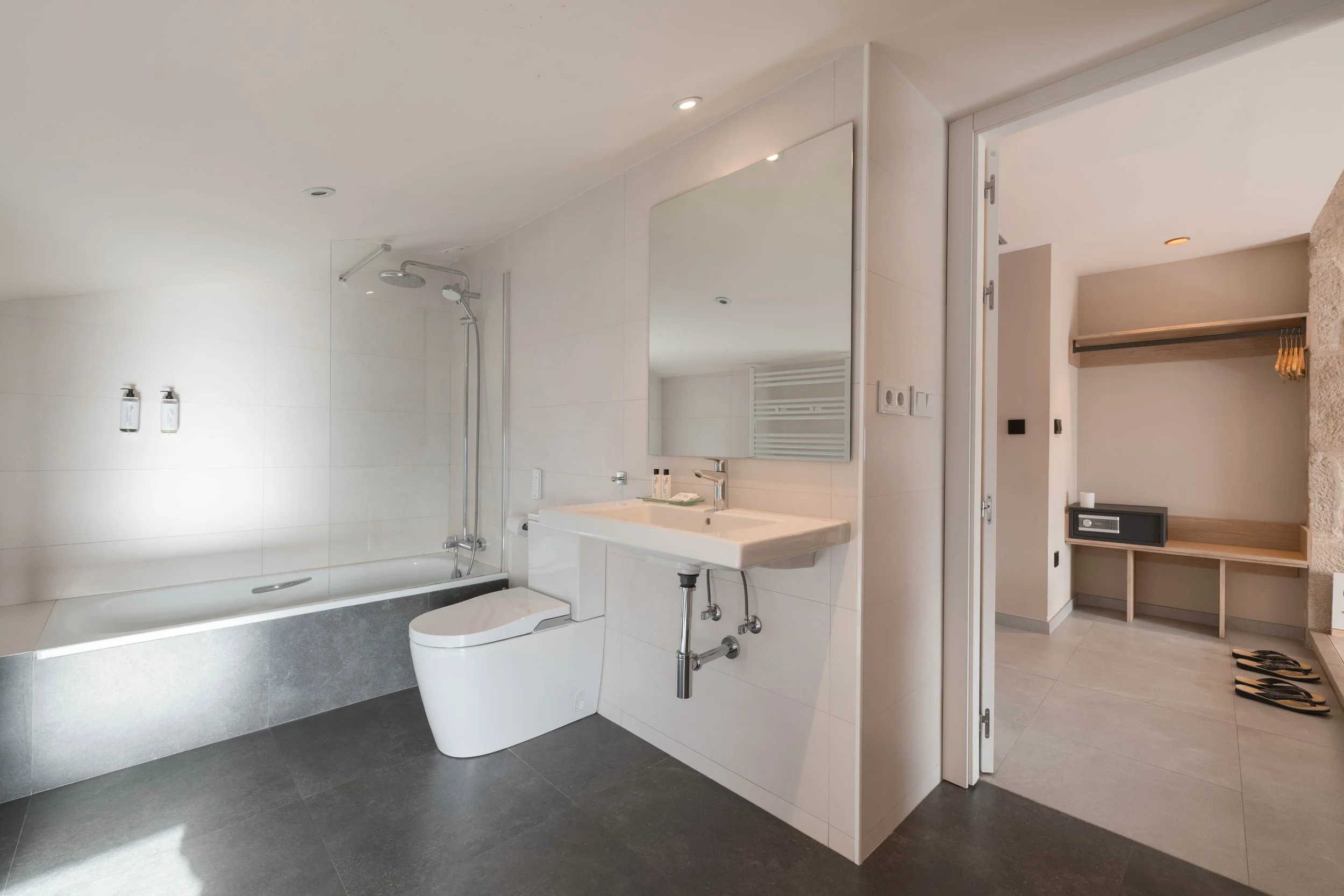 Baño moderno con ducha, inodoro y lavabo con espejo grande, habitación con armario y caja fuerte, pisos oscuros y paredes blancas.