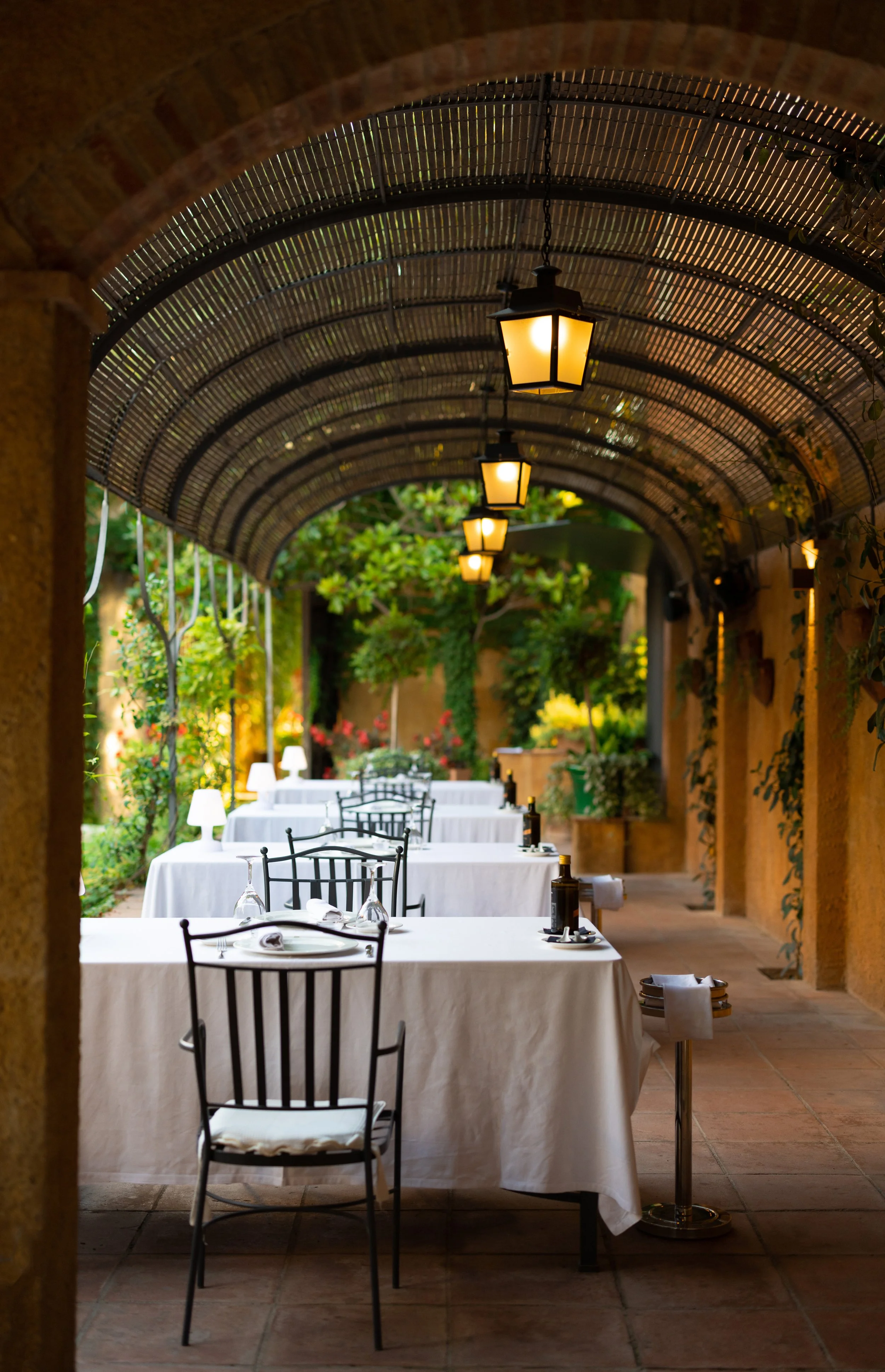 Comedor con mesas vestidas con manteles blancos y sillas de hierro, en un espacio cubierto con arcos y luces cálidas, rodeado de vegetación y plantas decorativas.