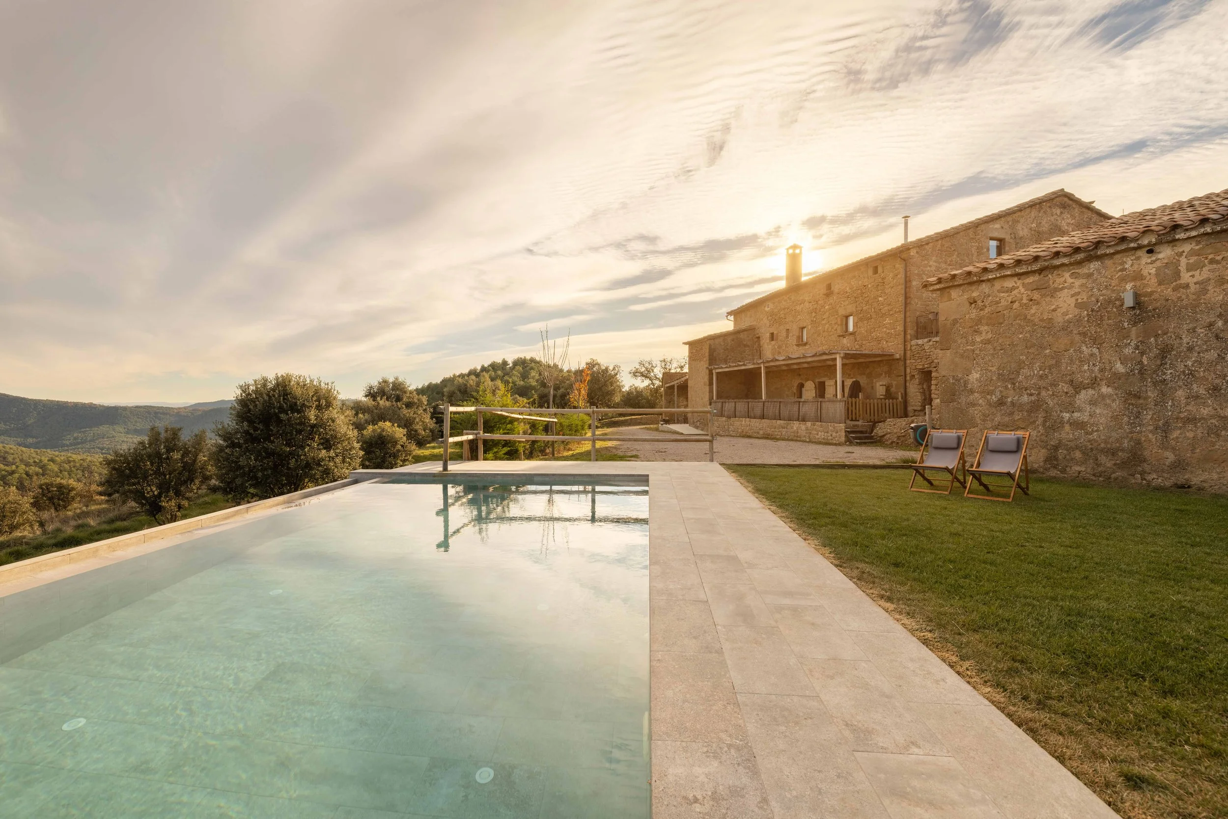 Vista de una piscina al atardecer junto a una casa de piedra con terraza, en un entorno rural con árboles y montañas en el fondo.
