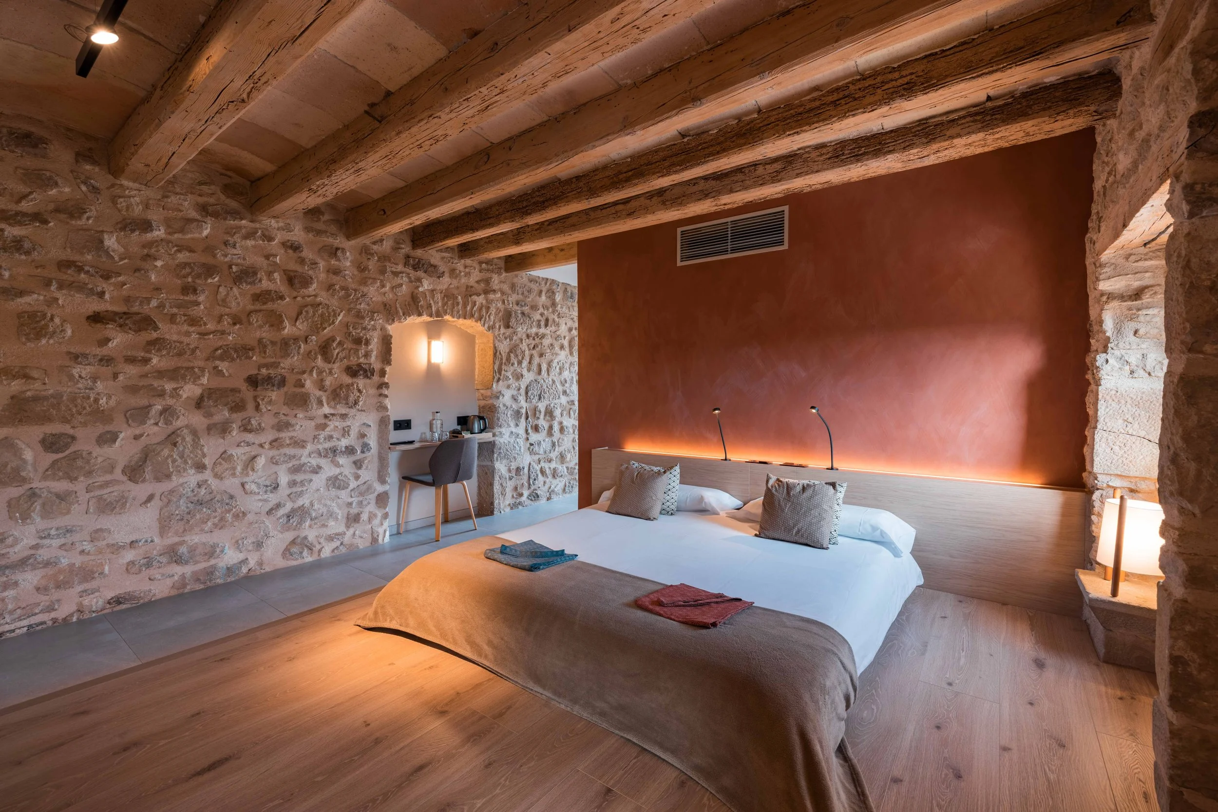 Habitación de hotel con paredes de piedra, techo de vigas de madera, cama con sábanas blancas y almohadas decorativas, lámparas de lectura, y área de trabajo con silla y máquina de café.