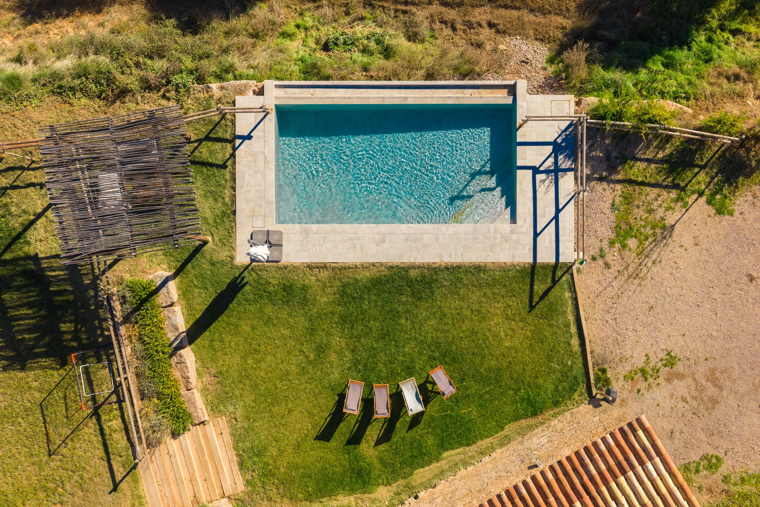Vista aérea de una piscina rectangular en un patio con césped, camastros, pérgola de madera, áreas de césped y tierra, y un tejado de tejas.