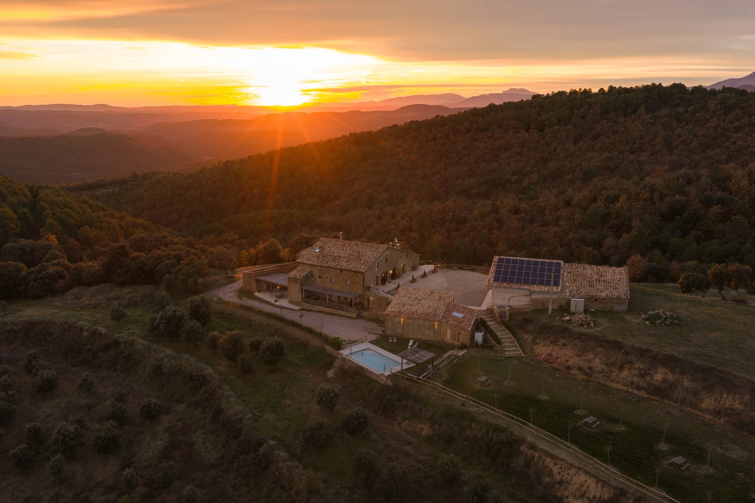 Casa de campo con piscina al aire libre y paneles solares, situada en un paisaje montañoso durante un atardecer.