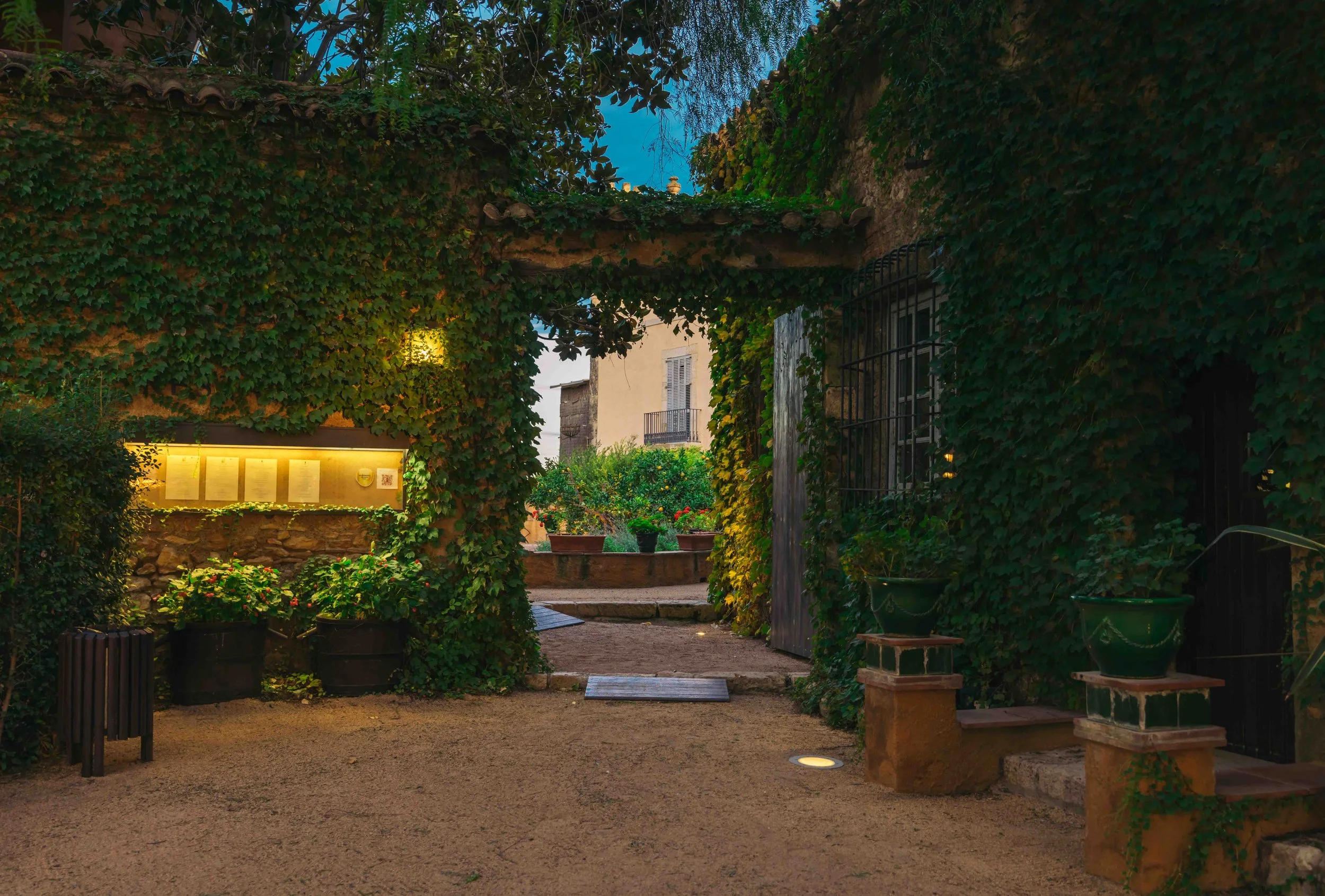 Jardín con entrada de arcos cubiertos de hiedra y plantas en macetas, con casas antiguas de fondo, de noche.