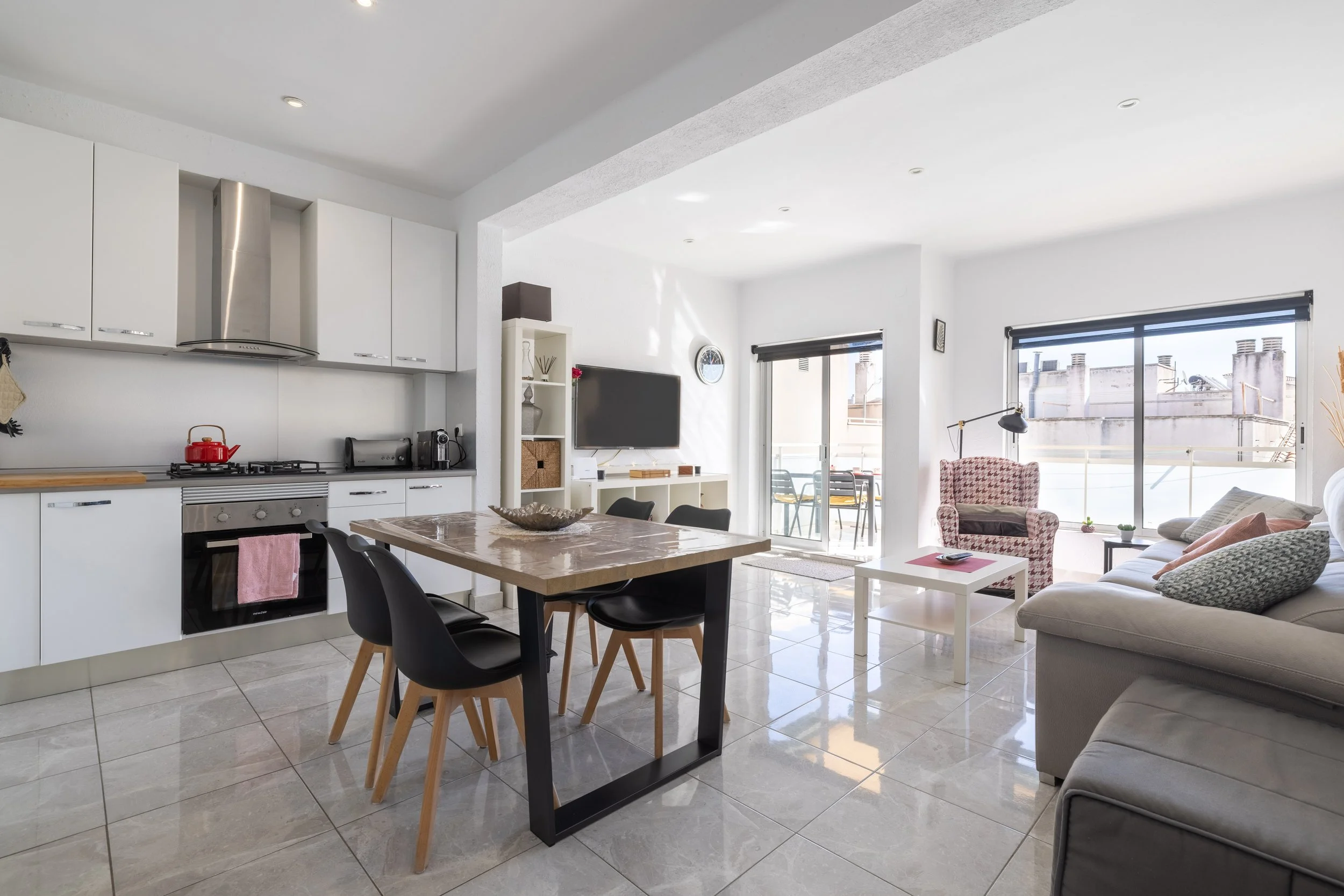 Sala de estar y cocina moderna con ventanas grandes, muebles blancos y un comedor con sillas negras de madera, perro durmiendo en un sillón gris y una mesa blanca.