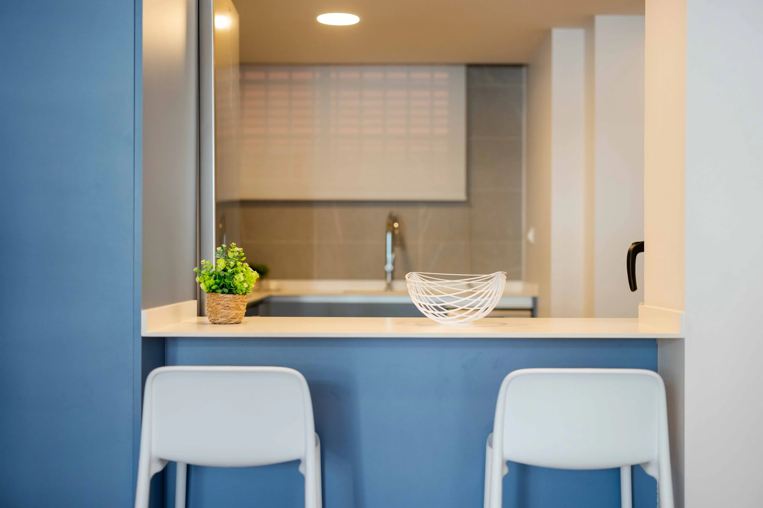 Vista de una cocina moderna con barra, dos sillas blancas delante, planta decorativa y una fuente de fruta sobre la barra, en colores azul, blanco y beige.