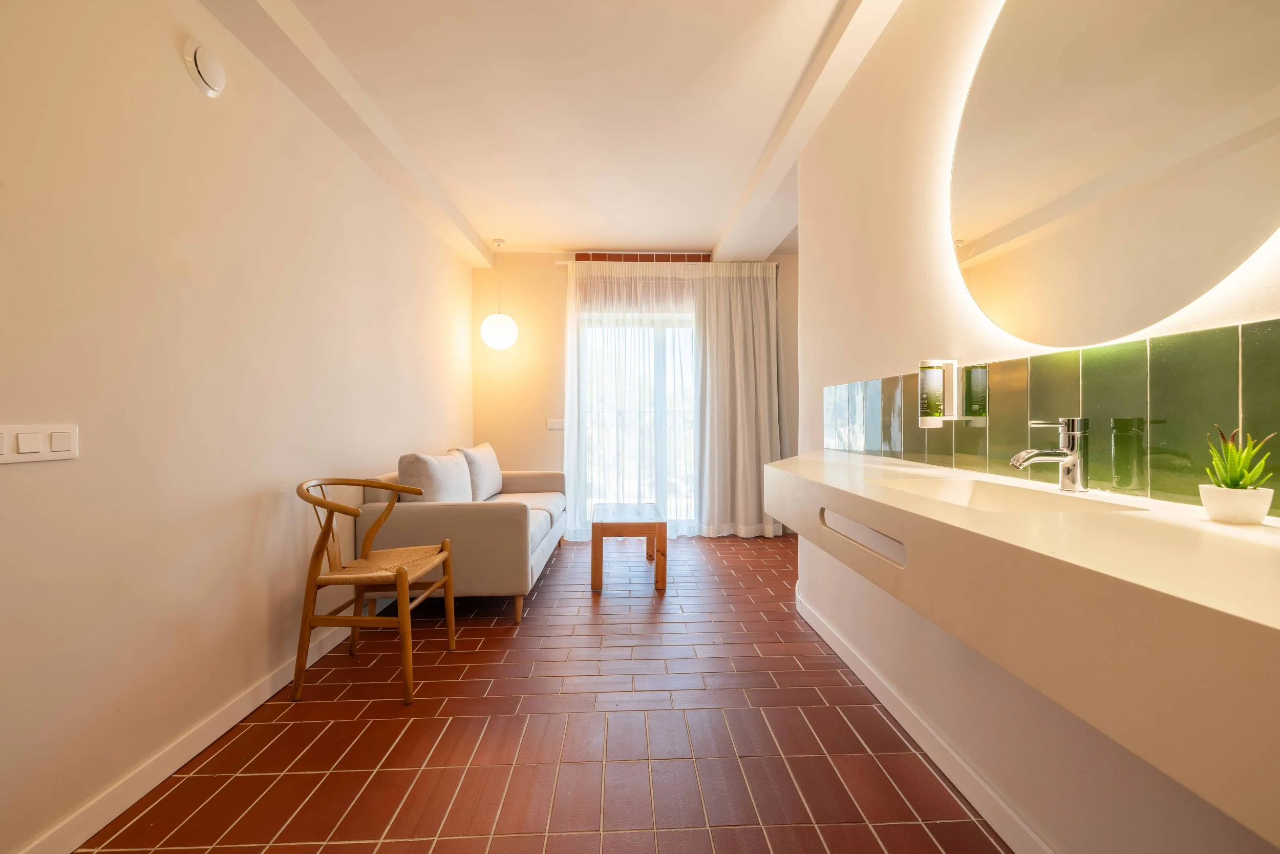 Sala de estar con sofá beige, silla de madera, mesa pequeña, cortinas blancas y espejo redondo en la pared; suelo de baldosas rojas.