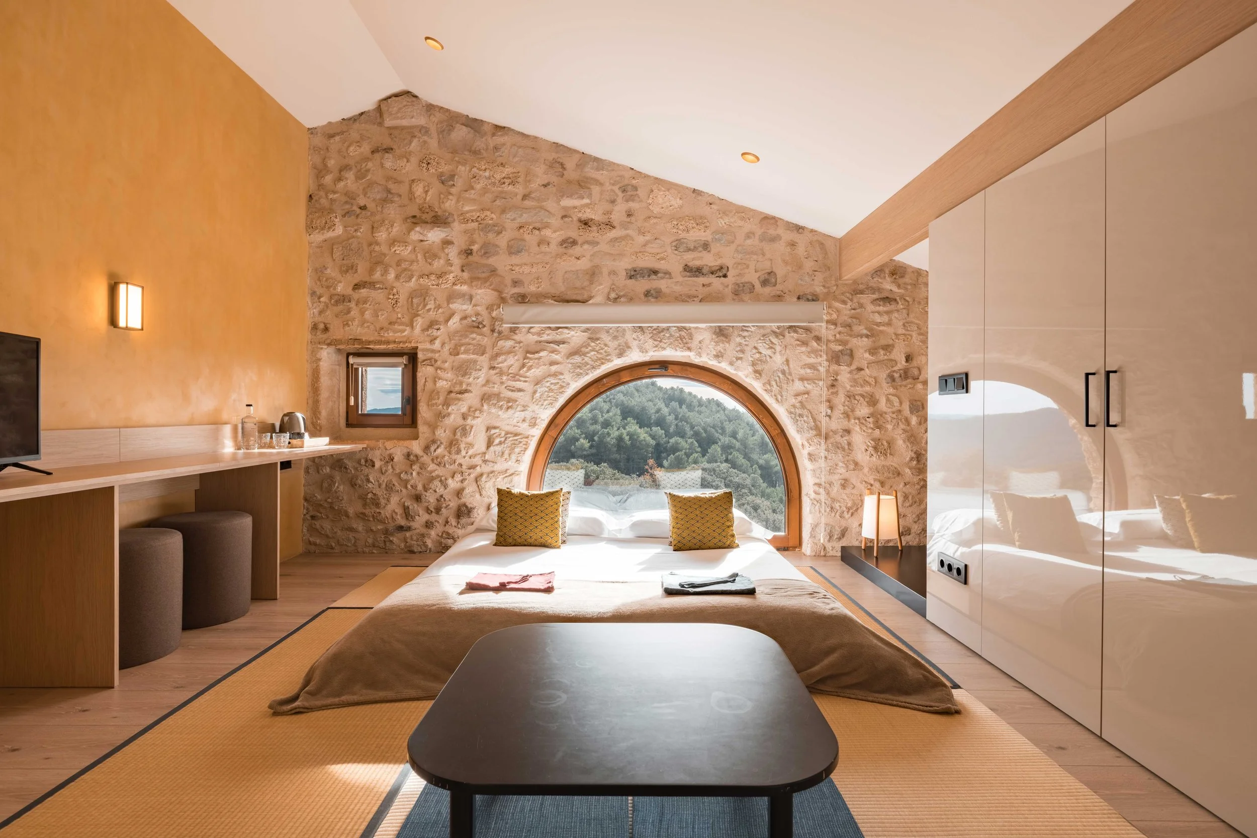 Habitación moderna con pared de piedra, cama doble con almohadas amarillas, ventana redonda con vista a árboles, mesa negra al pie de la cama, iluminación cálida, armario blanco grande, y decoración minimalista.