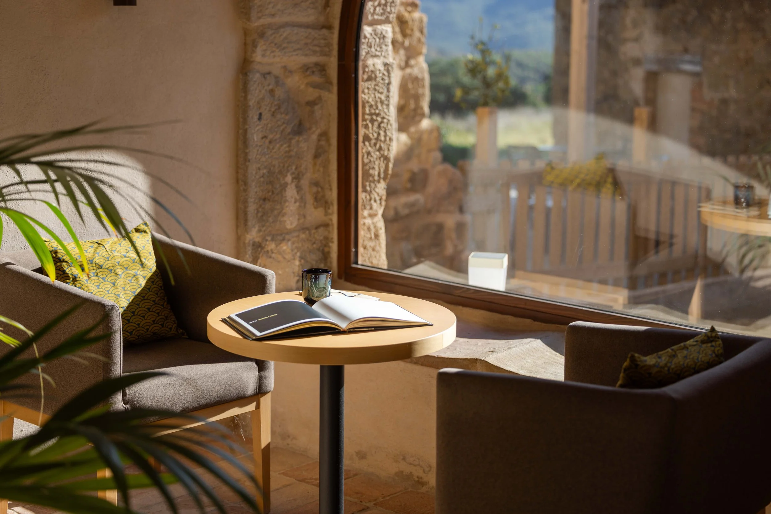 Interior de una sala con sillones grises, cojines decorativos, una mesa redonda de madera con un libro abierto y una taza, junto a una gran ventana que deja entrar luz natural y ofrece vista a un paisaje rural con árboles y una terraza de madera.