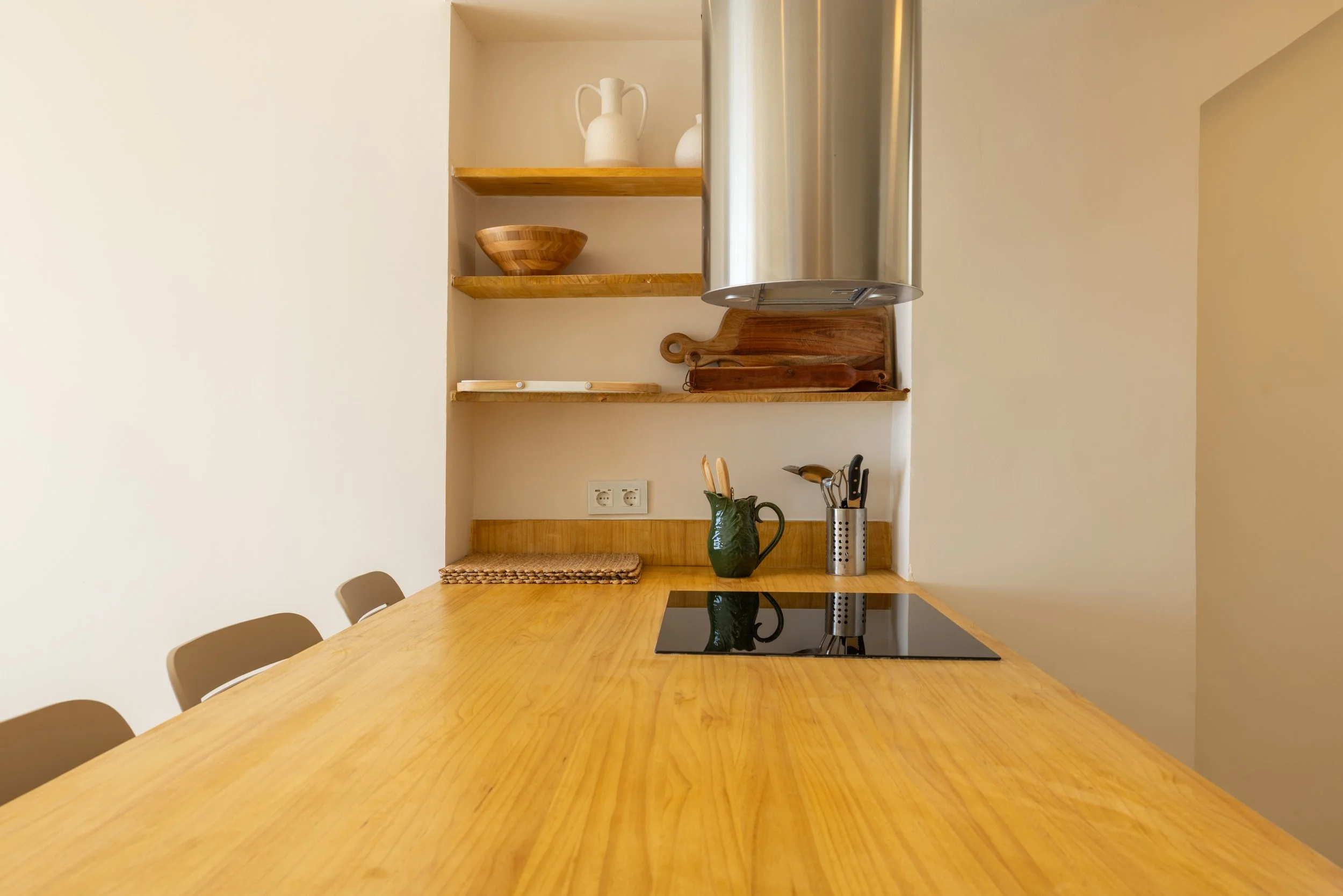 Encimera de madera con utensilios de cocina, incluyendo un cuchillo, un tenedor y una espátula en un recipiente de cerámica, con una estufa eléctrica y estantes con vajilla y accesorios de madera.