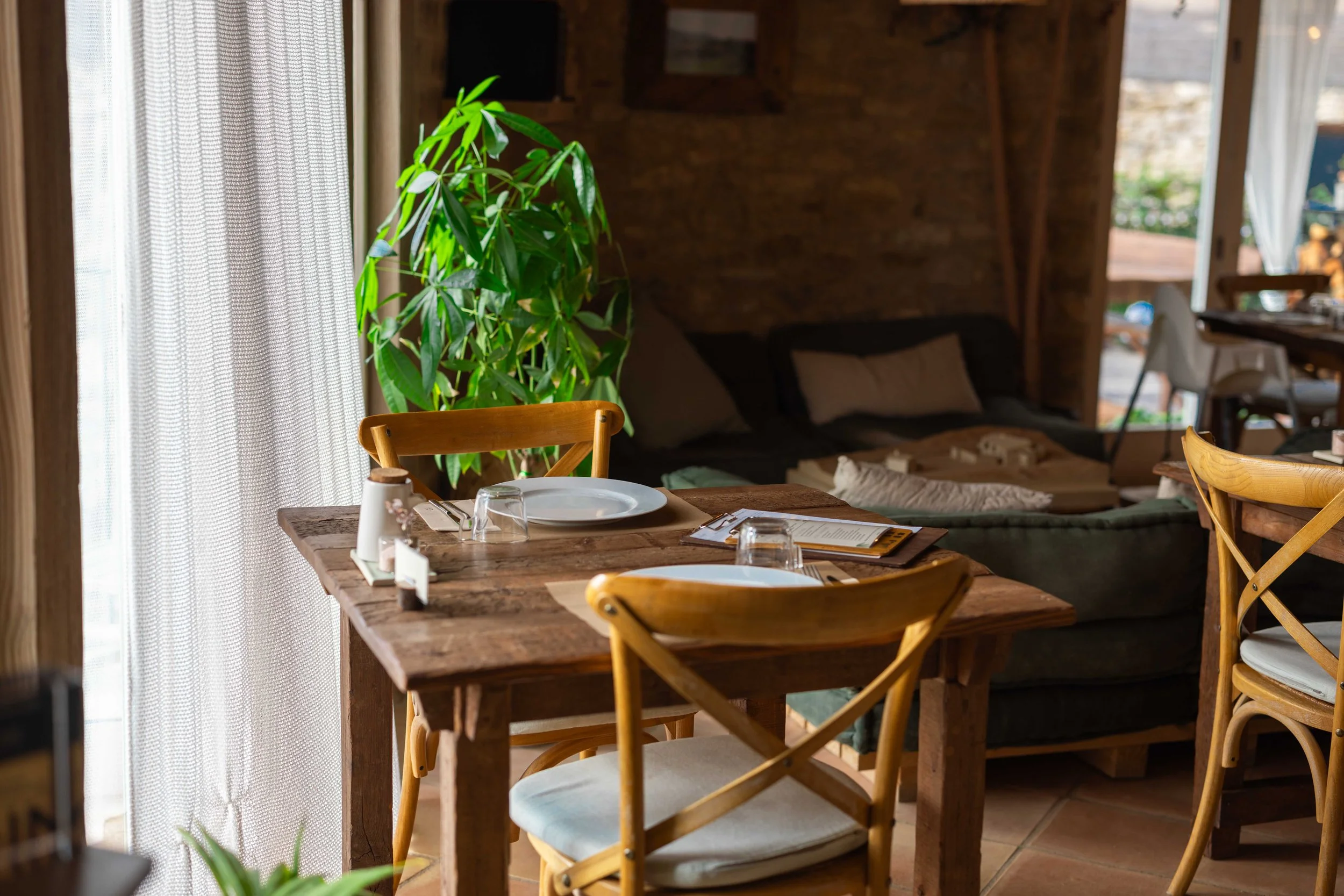Fotografía  de mesa de comedor en un ambiente rústico con platos, gafas y menús, cerca de una planta y una ventana con cortinas blancas.