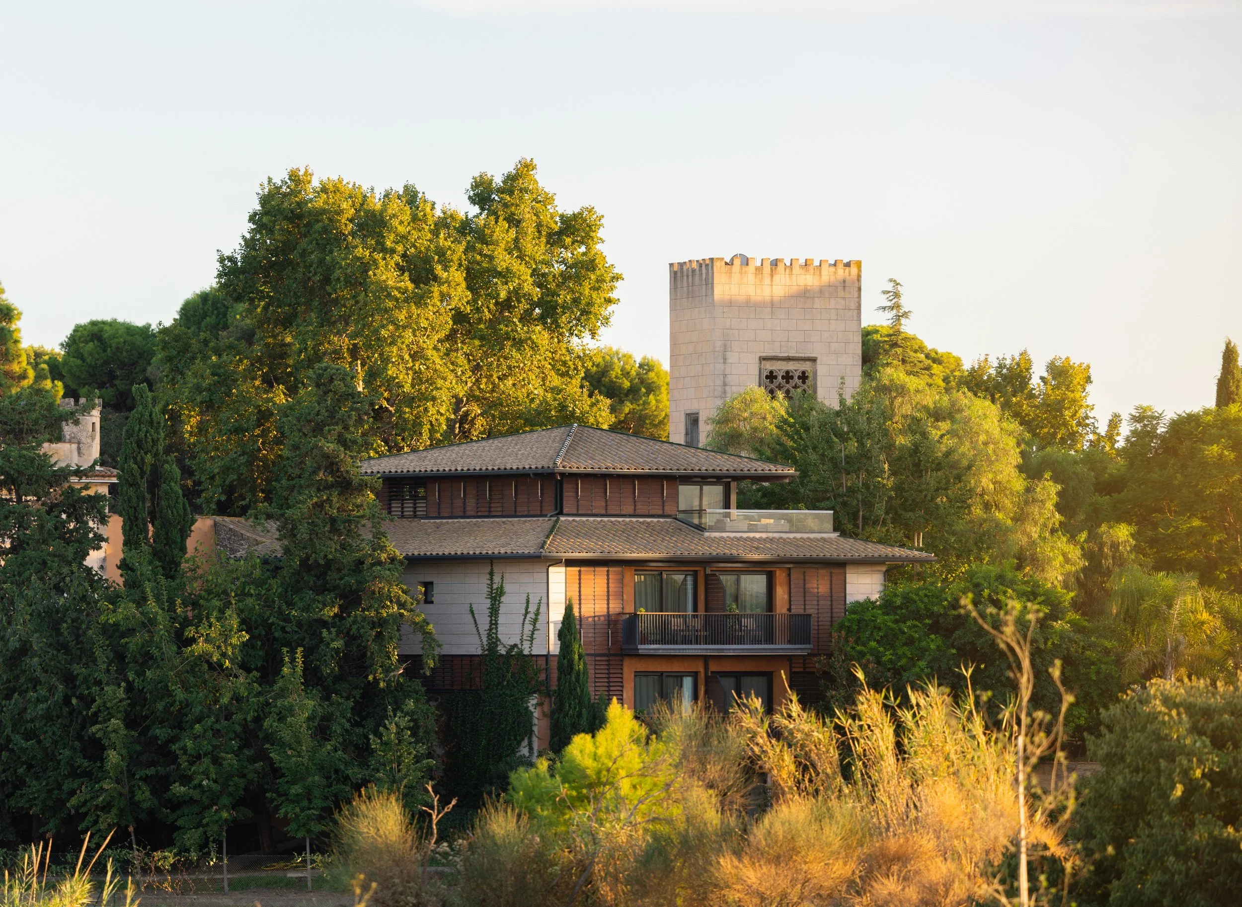 Casa moderna rodeada de árboles verdes y vegetación, con una torre en el fondo y luz de atardecer.