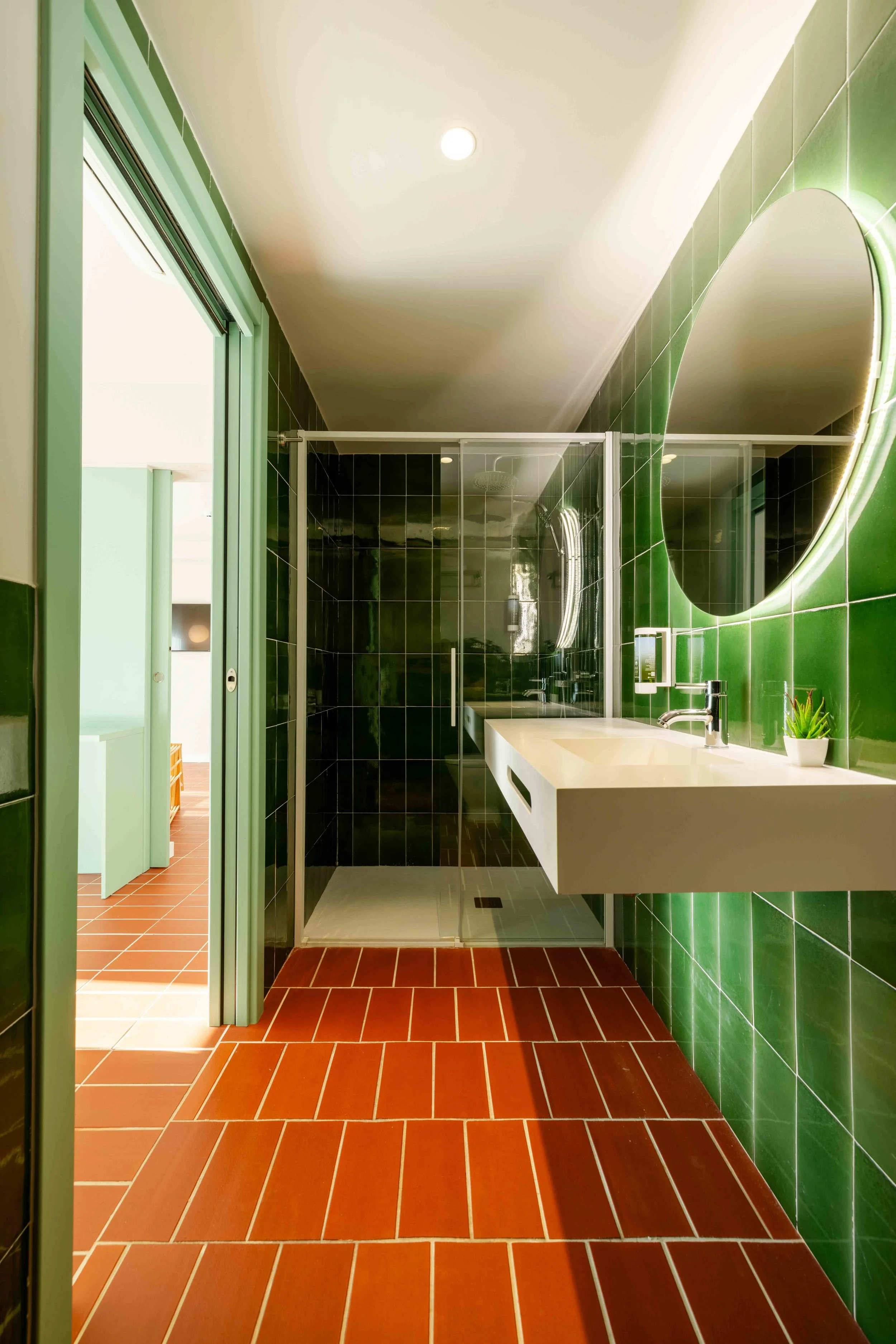 Baño con paredes verdes, piso de baldosas rojas, y una ducha con puerta de vidrio, lavabo blanco flotante y espejo encendido, vista desde la entrada