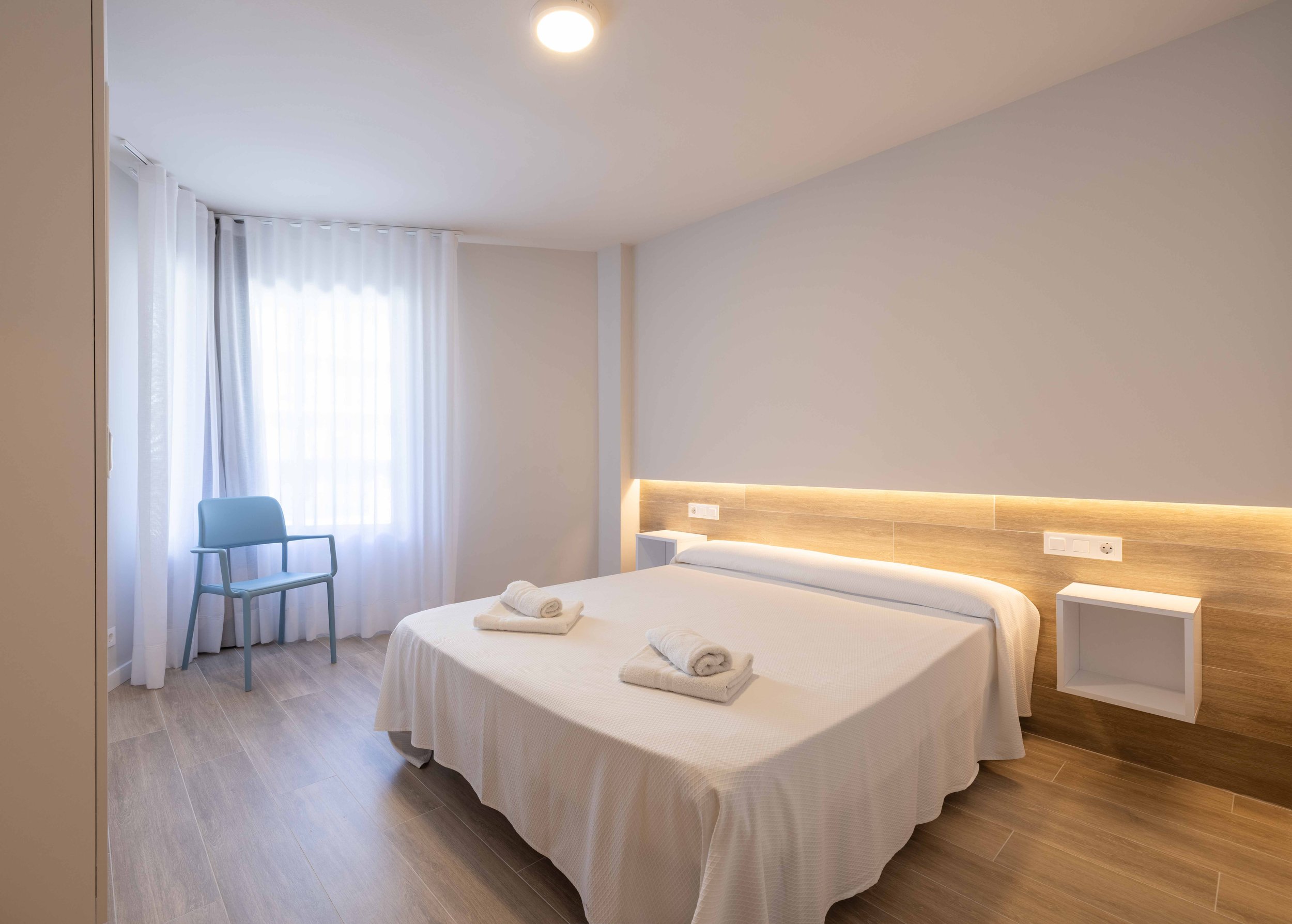 Habitación de hotel minimalista con cama doble, toallas dobladas en la cama, sillón azul, cortinas blancas, paredes beige y suelo de madera.