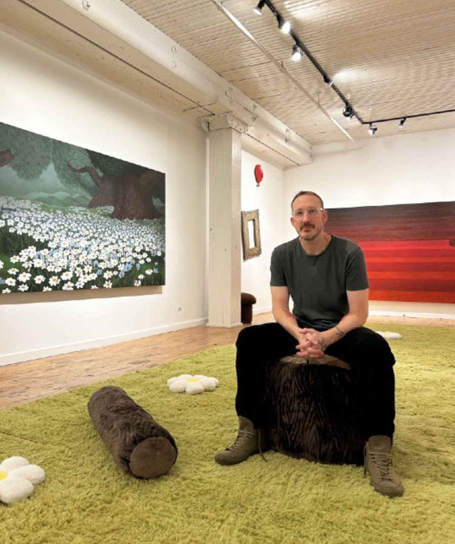 A man sitting on a furry black stool inside an art gallery, surrounded by a green rug, with artworks on the walls including a large painting of a tree with a field of white flowers, and a red wall panel. Decorative plush flowers and a brown log-shaped object are on the rug.