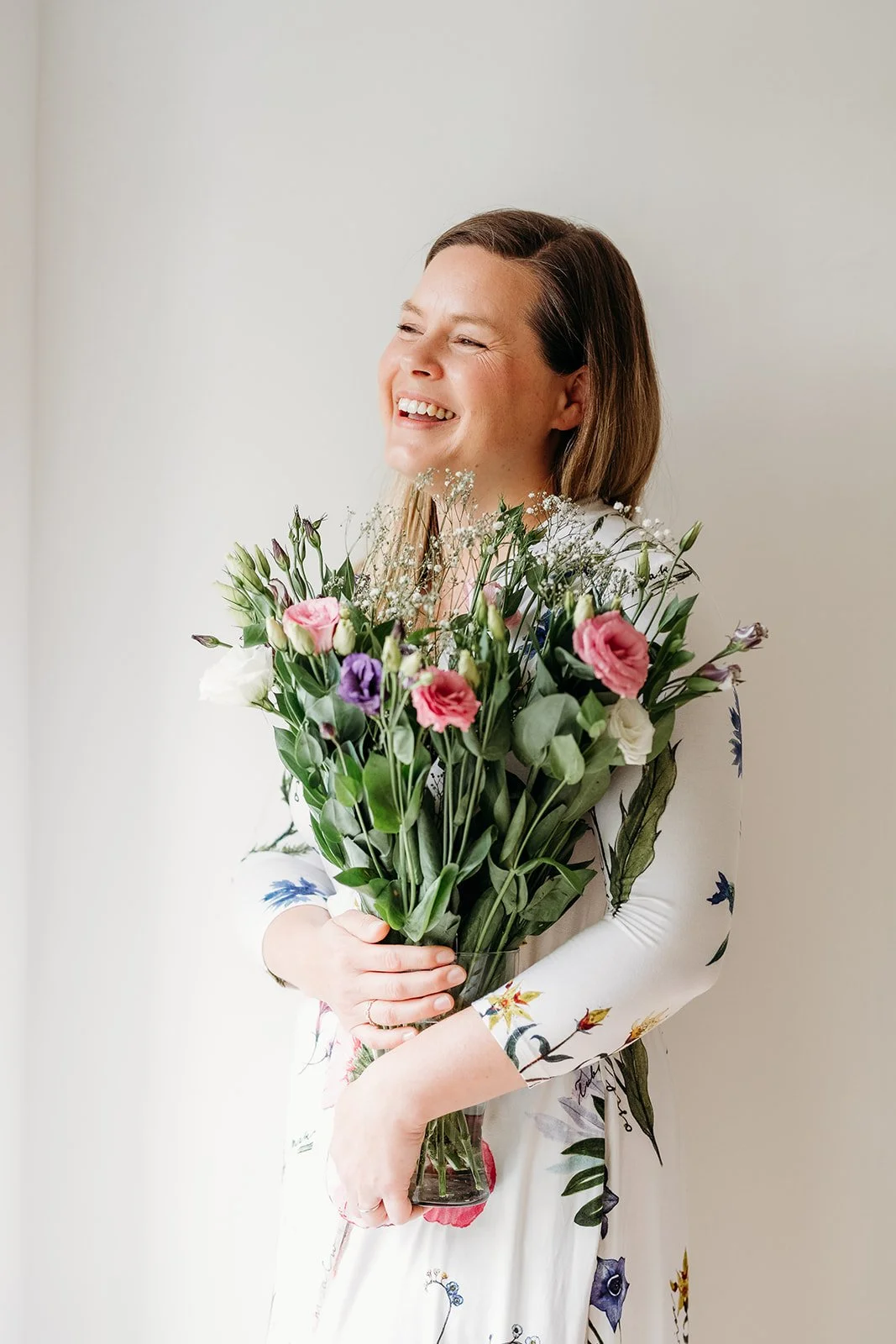 A woman smiling and holding a large bouquet of colorful flowers, standing against a plain white wall. Surrey-based Flying Star Feng Shui practitioner helping clients globally navigate big life transitions. Sell, buy, and transform your home into a sp