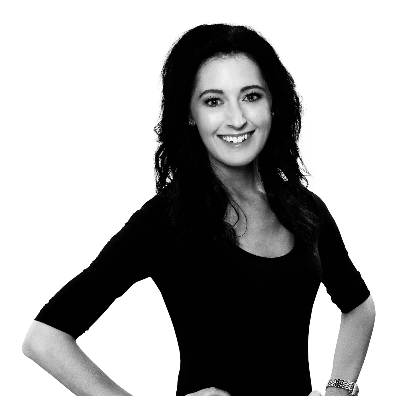 Black and white portrait of a smiling woman with dark, wavy hair, wearing a black top and a watch on her left wrist, standing with her hands on her hips.