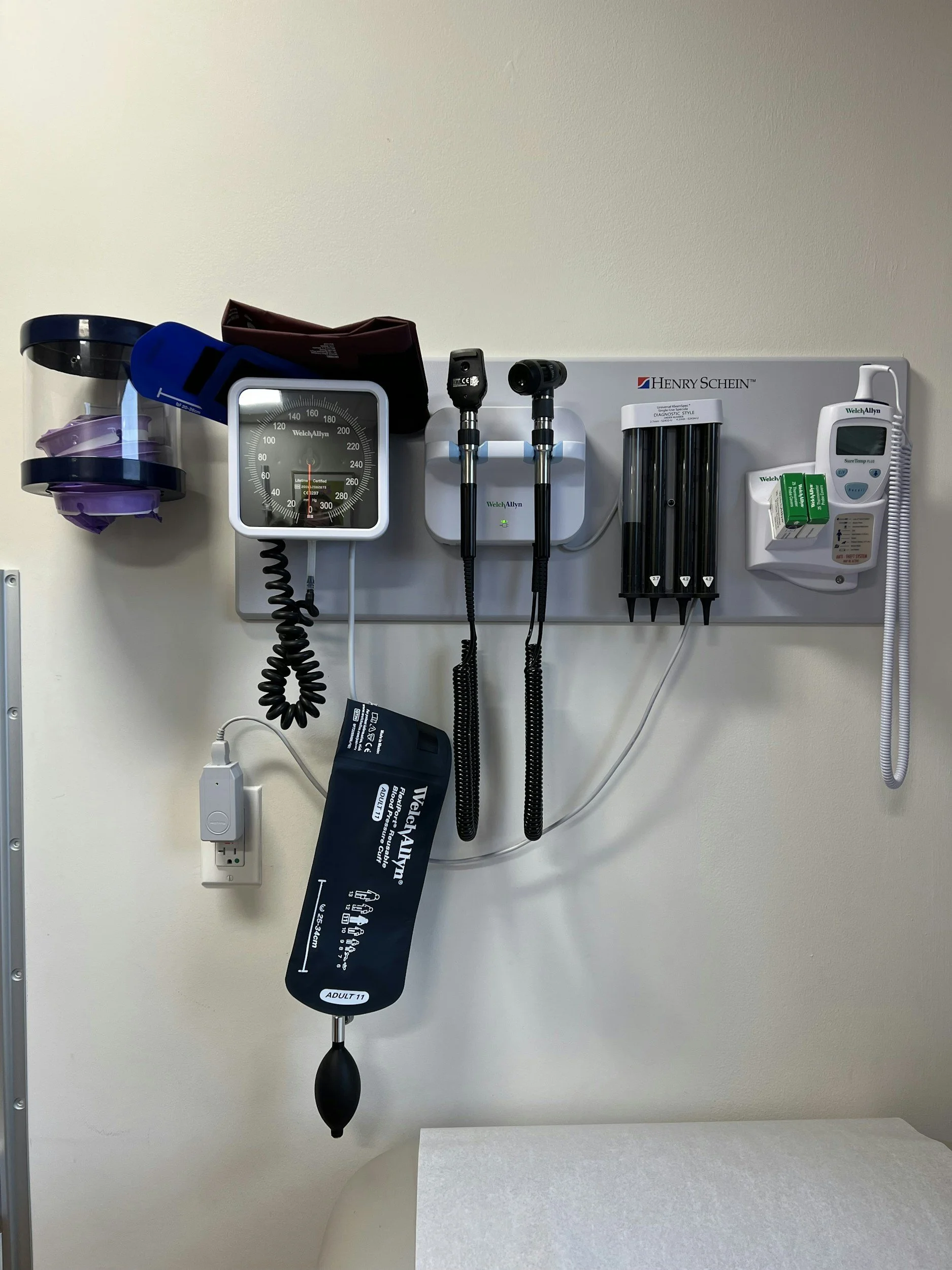 Medical equipment on a wall, including blood pressure monitor, otoscope, ophthalmoscope, and other diagnostic tools.