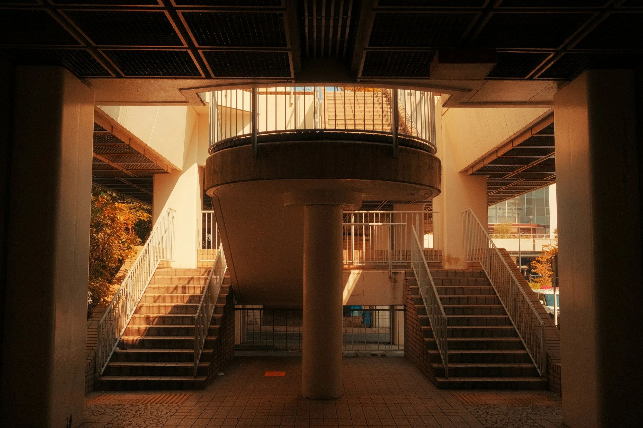 Underpass with stairs on both sides leading to an upper level, with sunlight illuminating the scene and a central concrete pillar supporting the structure.