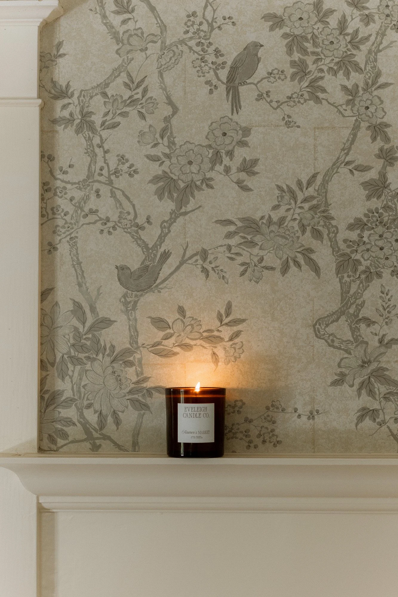 Lit candle on a mantel in front of aesthetic floral and bird patterned wallpaper. Home decor.