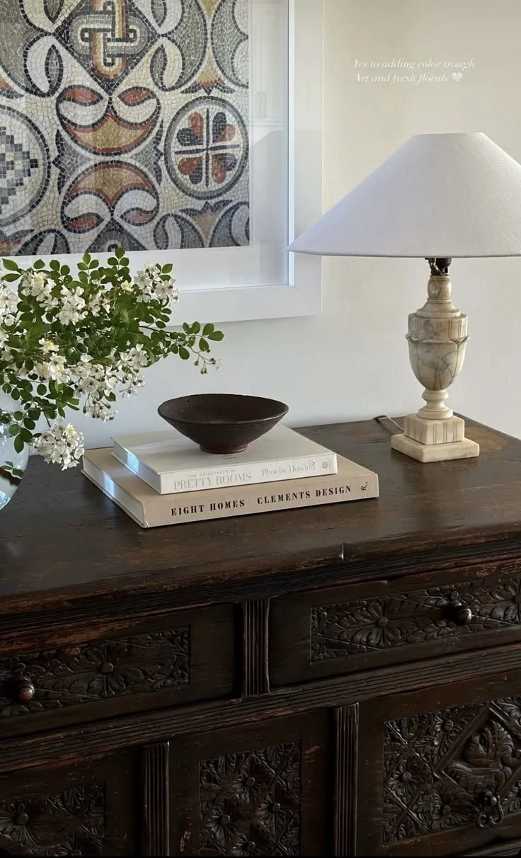 Room decor with a carved wooden table, books, a vase with white flowers, a ceramic bowl, a marble lamp, and a mosaic art piece on the wall.