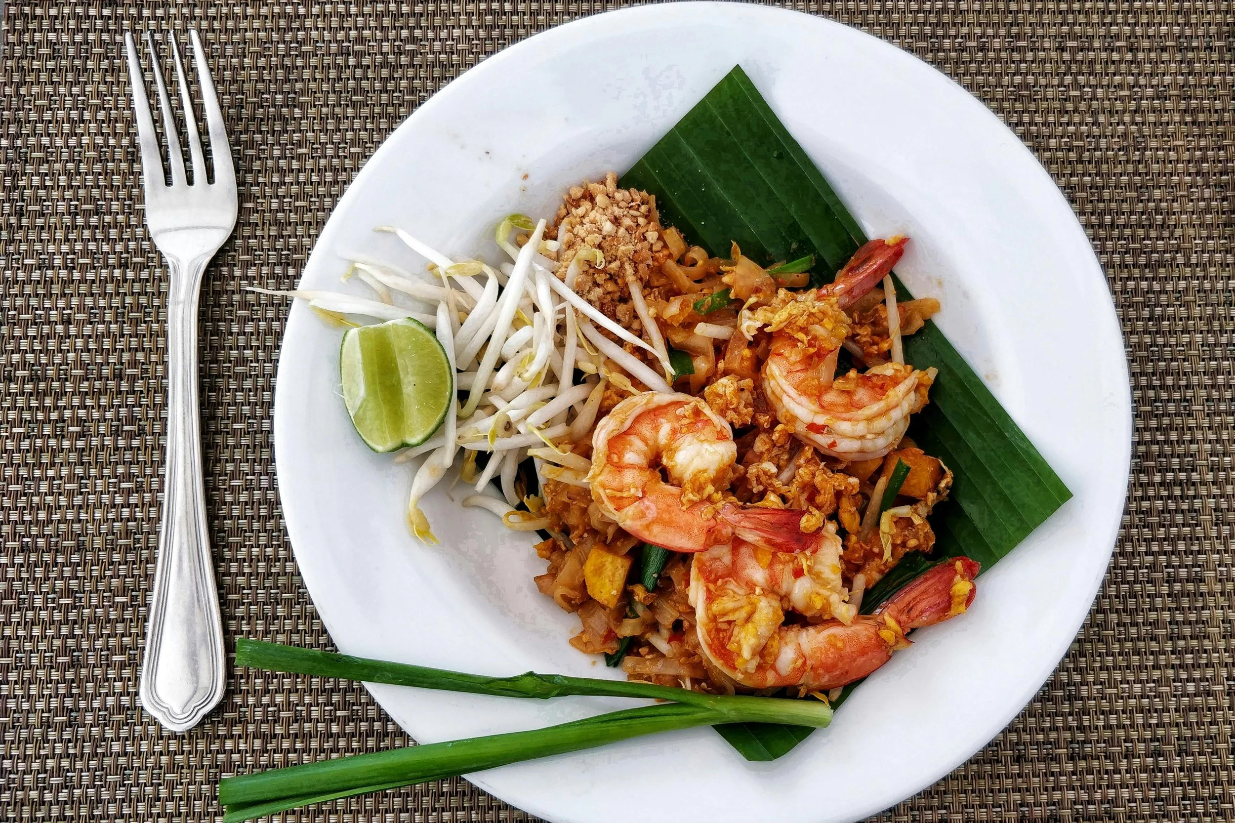 A white plate of Thai shrimp pad thai with lime wedge, bean sprouts, crushed peanuts, and green onion on a woven placemat with a fork beside it.