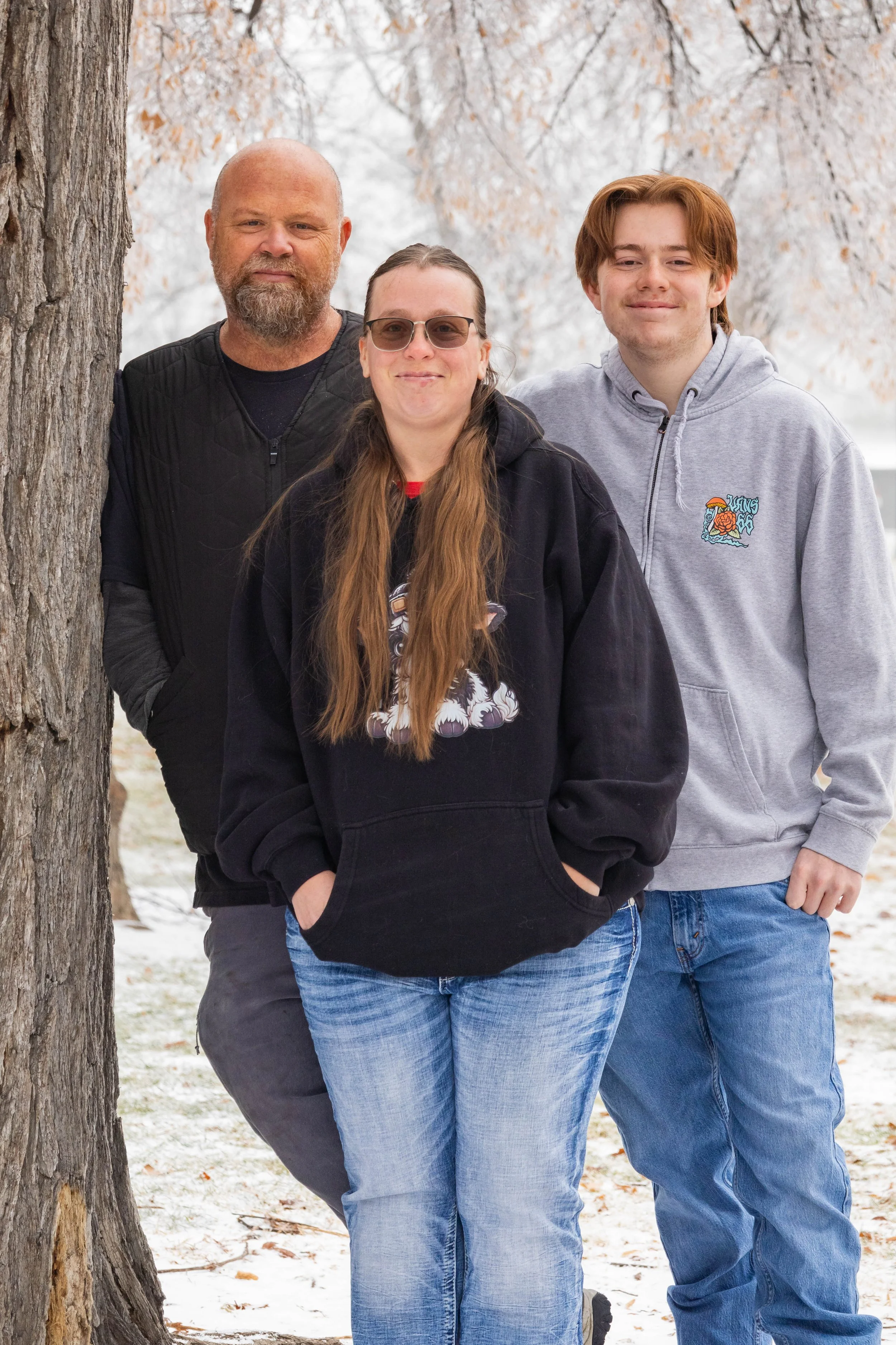 A family of three standing outdoors in winter, with bare trees and snow on the ground. The man has a beard and is wearing a black jacket, the woman has long hair and glasses, wearing a black hoodie, and the young man has red hair, wearing a gray hoodie with a logo, and all are smiling.